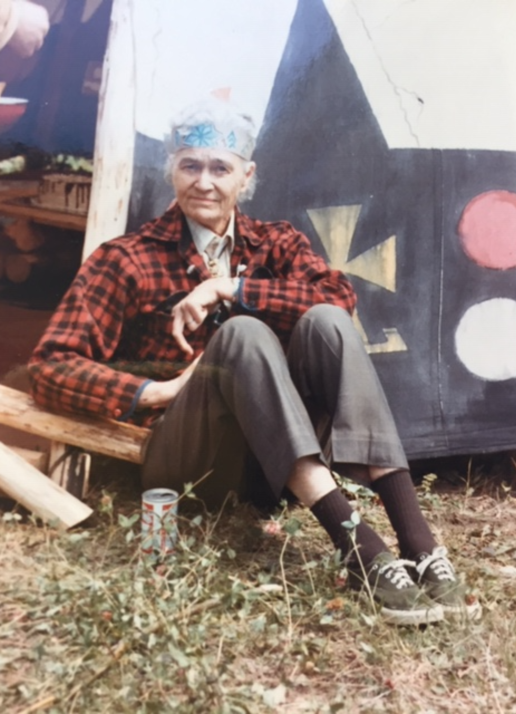 An elderly woman sitting on a wooden bench outdoors, wearing a plaid jacket, gray pants, and sneakers. She has a headband and is positioned in front of a painted backdrop with geometric shapes. A small can is on the ground next to her, and the background shows grass and outdoor elements.
