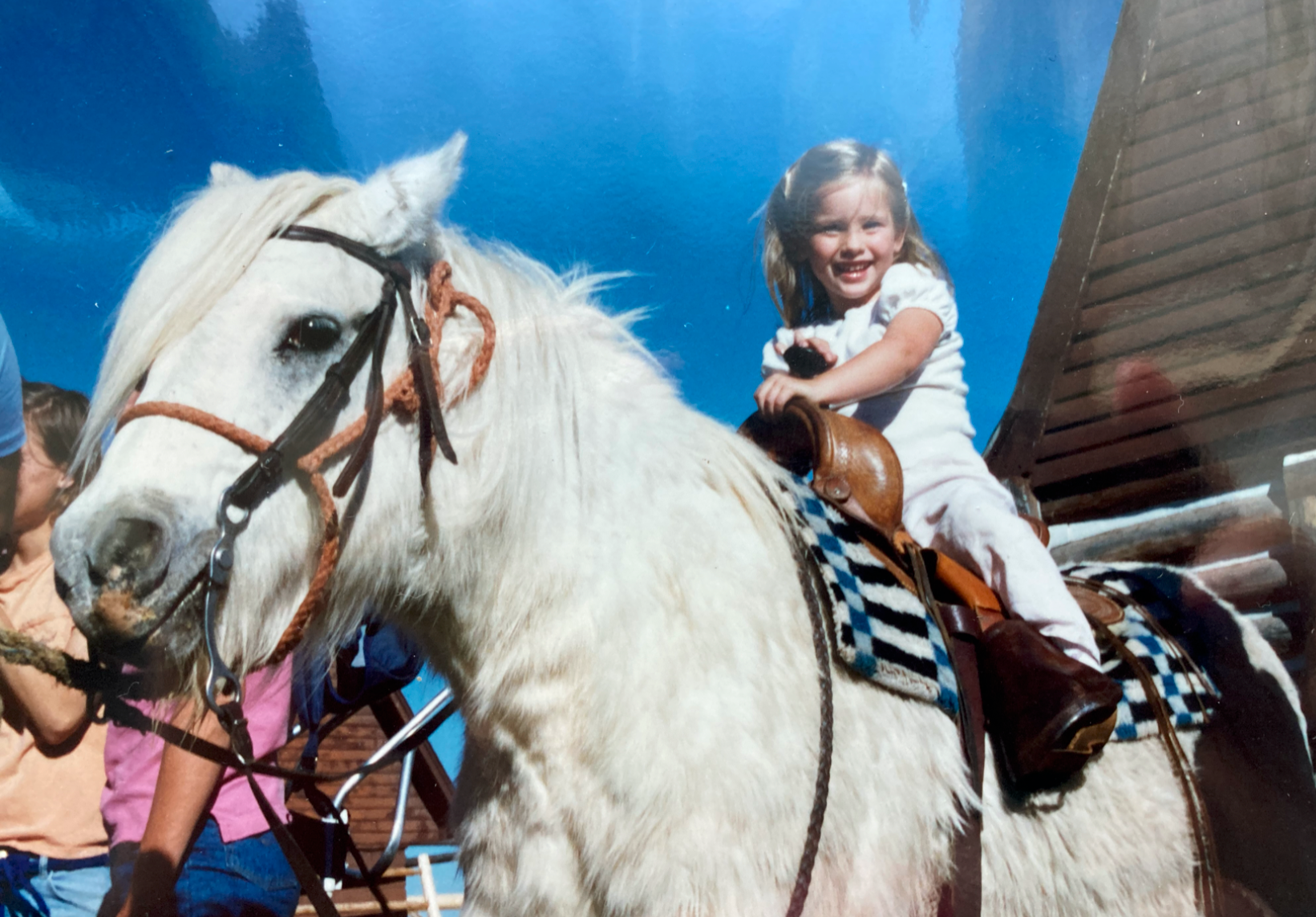 A young girl with a big smile riding a white horse outdoors on a sunny day, with a clear blue sky and part of a wooden building visible in the background.
