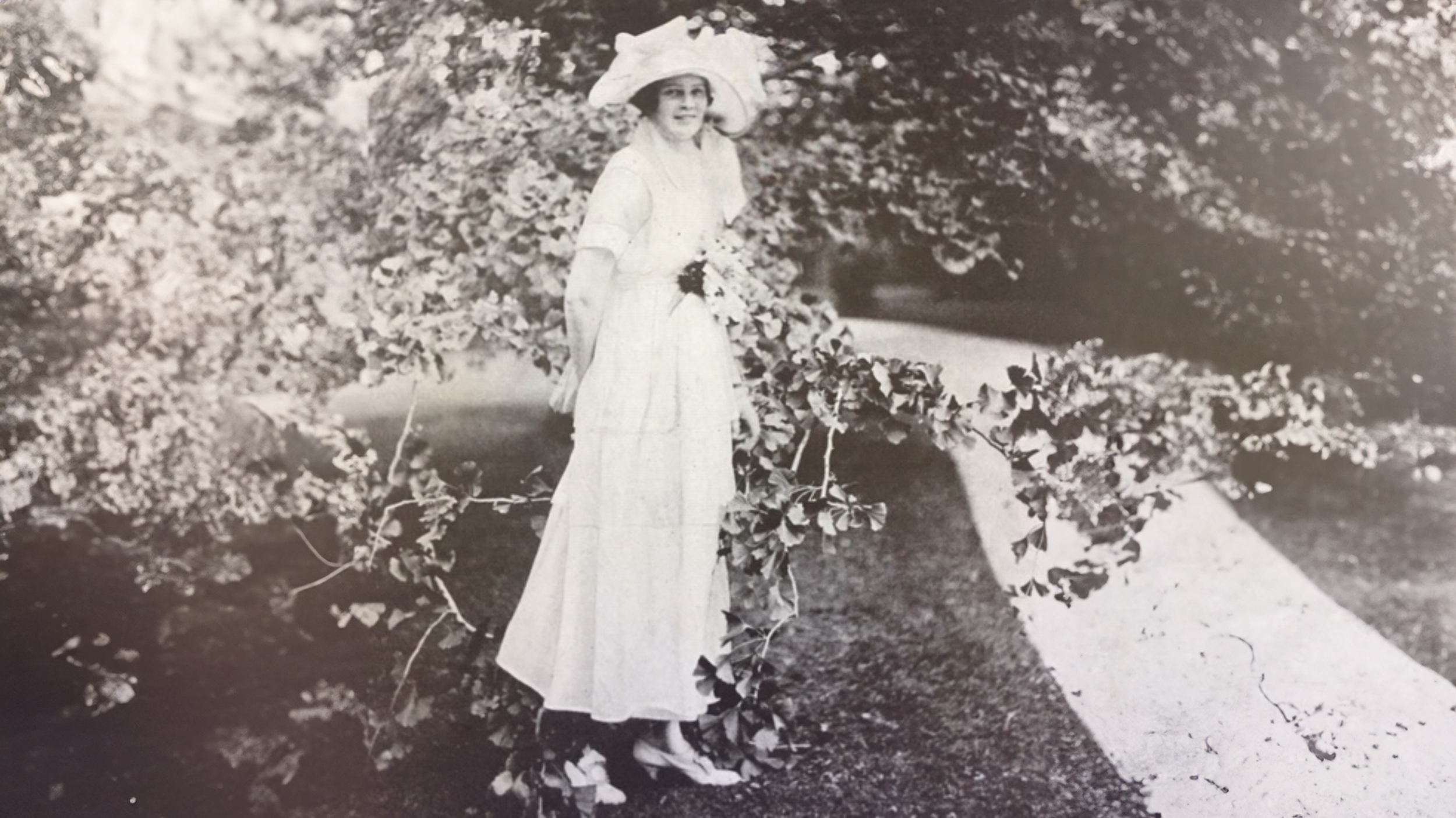 A woman in a long dress and wide-brimmed hat standing outdoors among bushes and trees.