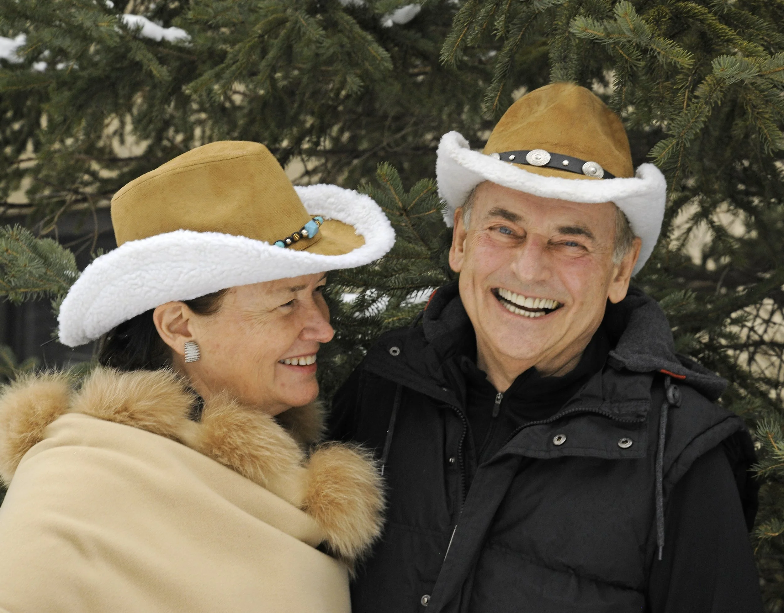 Older couple smiling and looking at each other, dressed warmly with cowboy hats, in front of a tree.