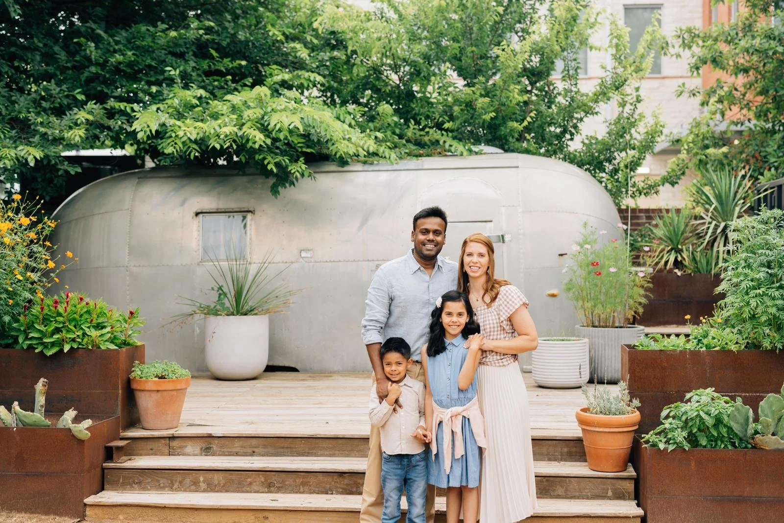 Family of four standing on a wooden deck in front of a silver Airstream trailer surrounded by potted plants and greenery.