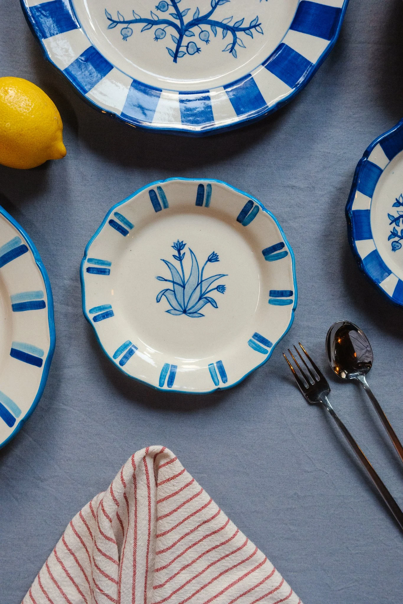 STUDIO PURANI Tulip dessert plate in blue styled with matching Pomegranate plates, a fork and spoon, and lemon — hand-painted blue and white ceramic tableware