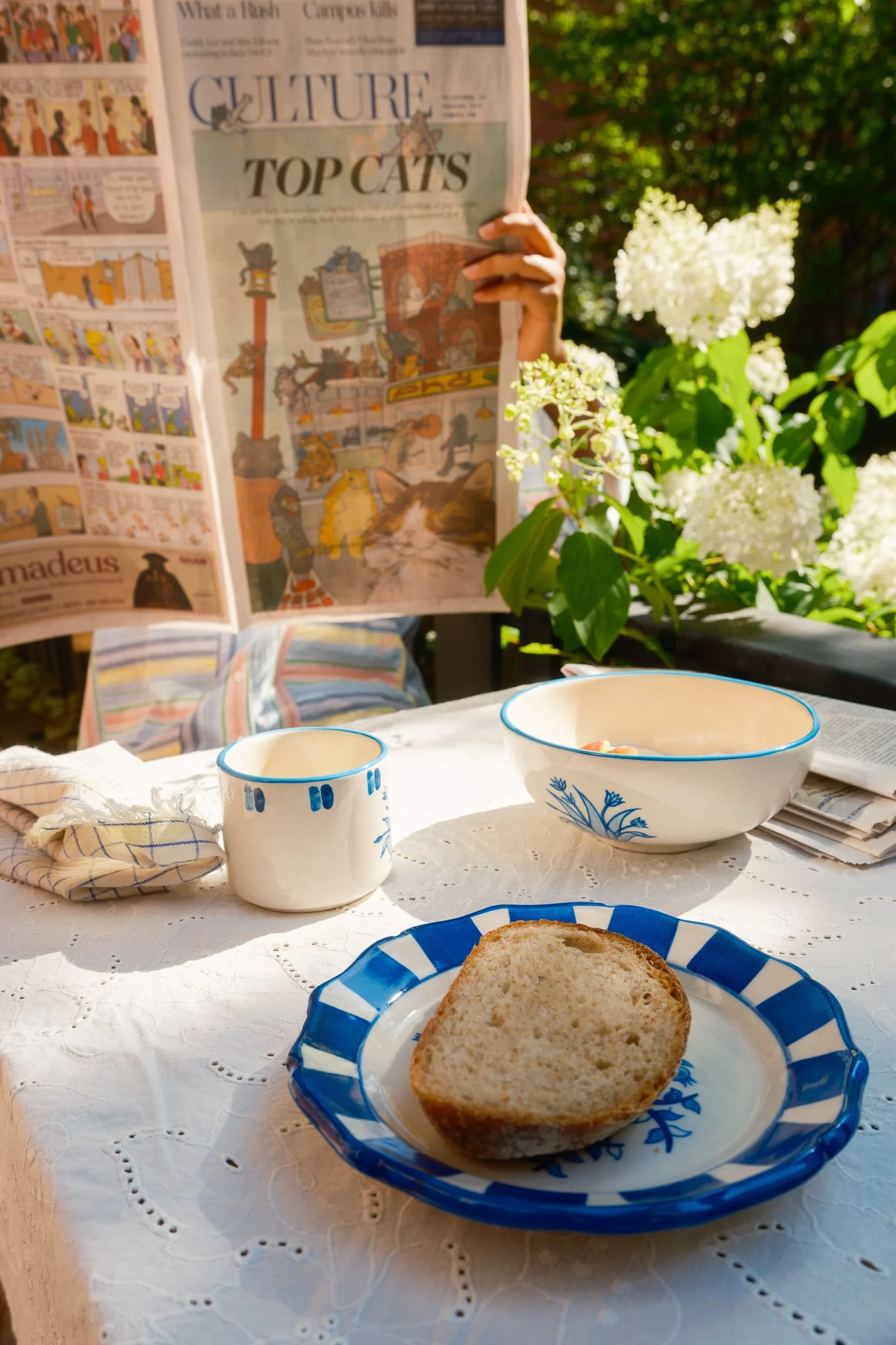 STUDIO PURANI Tulip breakfast bowl in blue styled outdoors with a matching Tulip espresso cup, pomegranate dessert plate, bread, and newspaper — hand-painted ceramic breakfast set