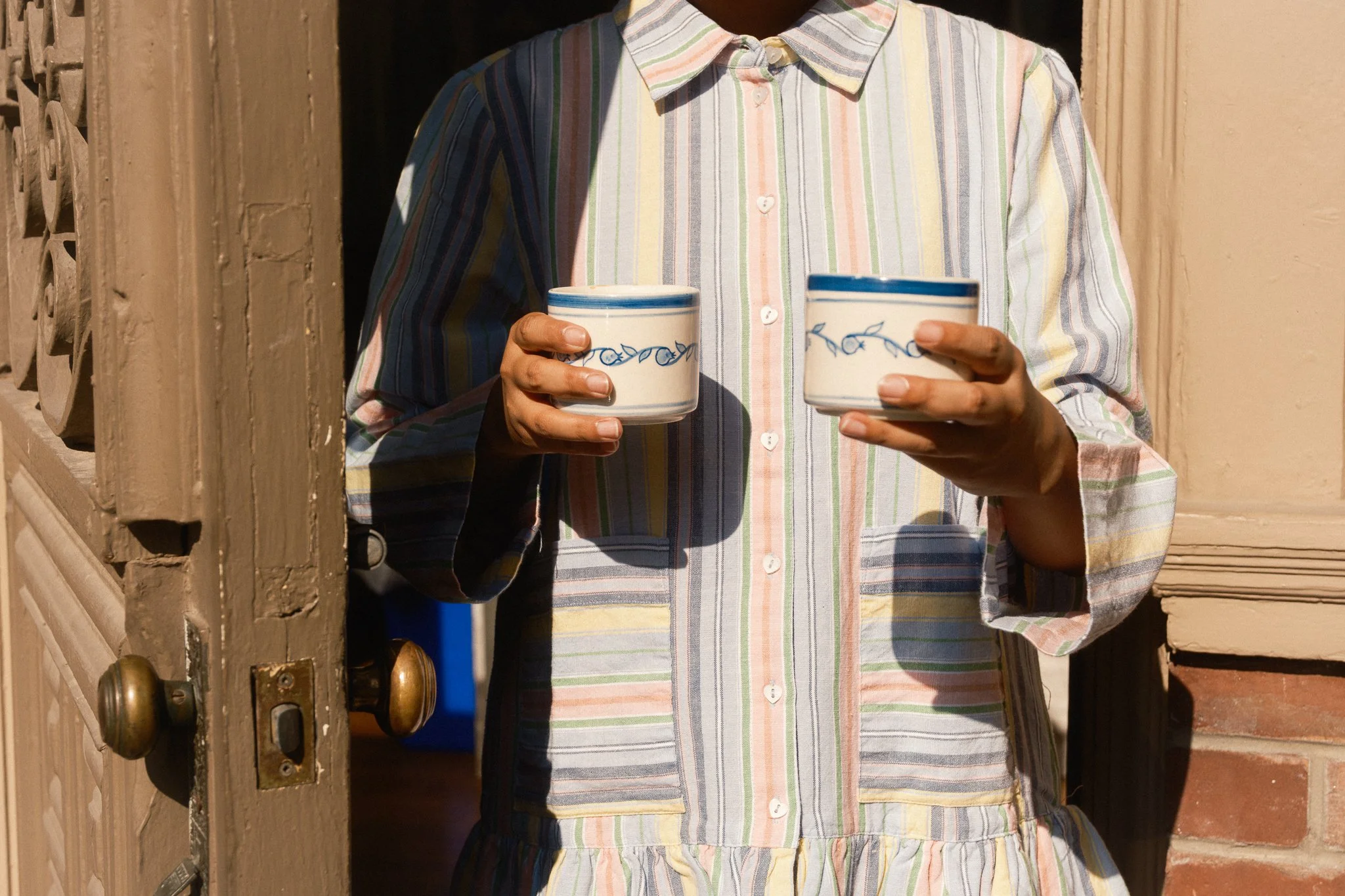 STUDIO PURANI Pomegranate cappuccino cups in blue held in two hands in front of a striped shirt — hand-painted ceramic cappuccino cups