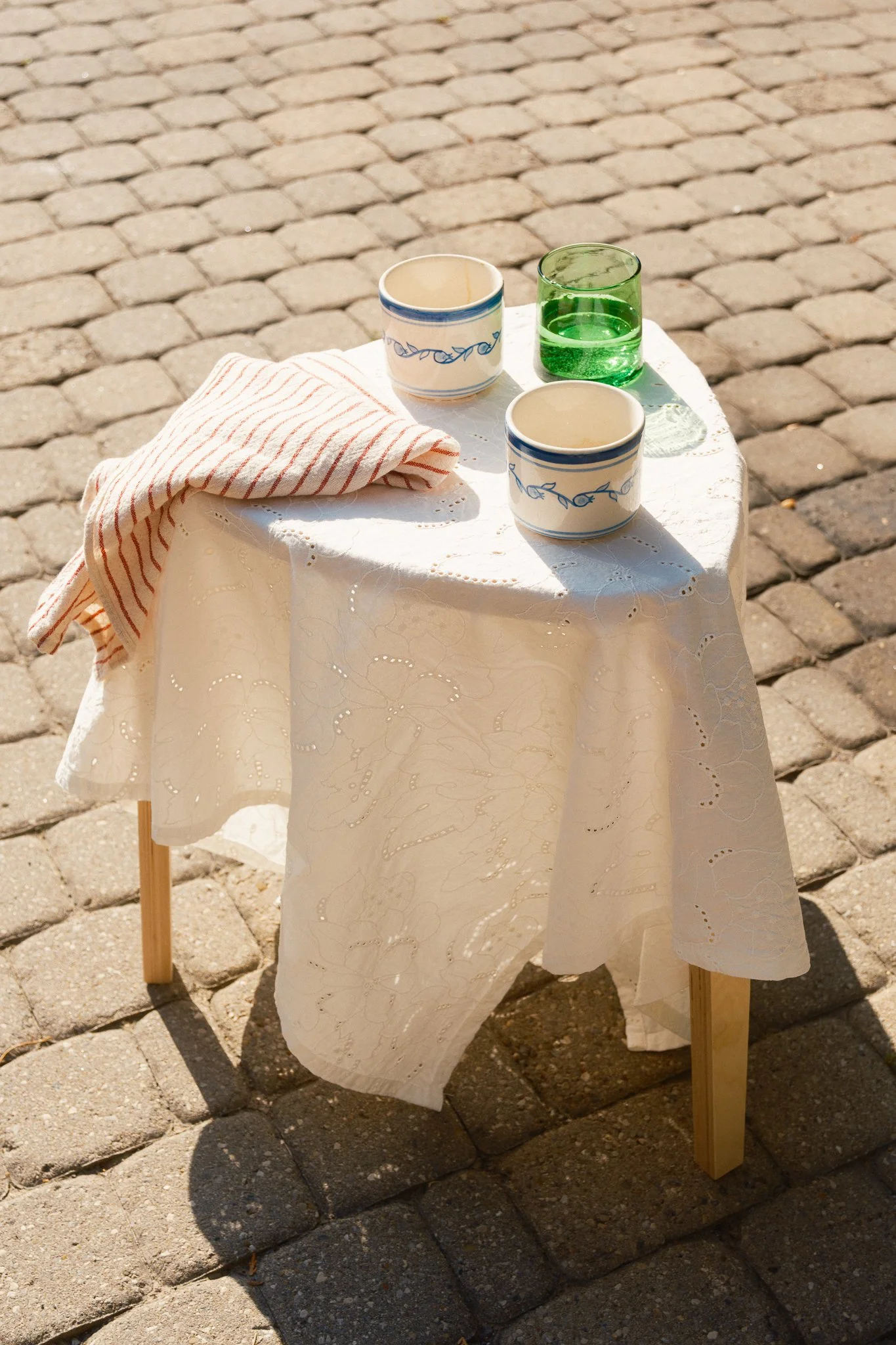 STUDIO PURANI Pomegranate cappuccino cups in blue on an outdoor side table with a green glass and striped linen napkin — hand-painted ceramic cappuccino cups styled outdoors