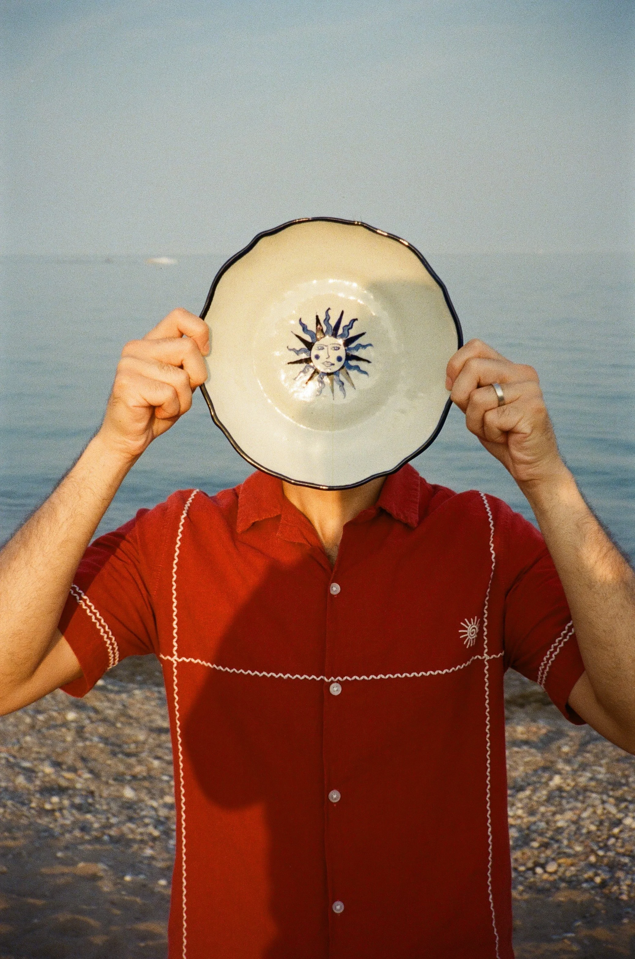 Person holding STUDIO PURANI plate with sun design in front of their face at a beach