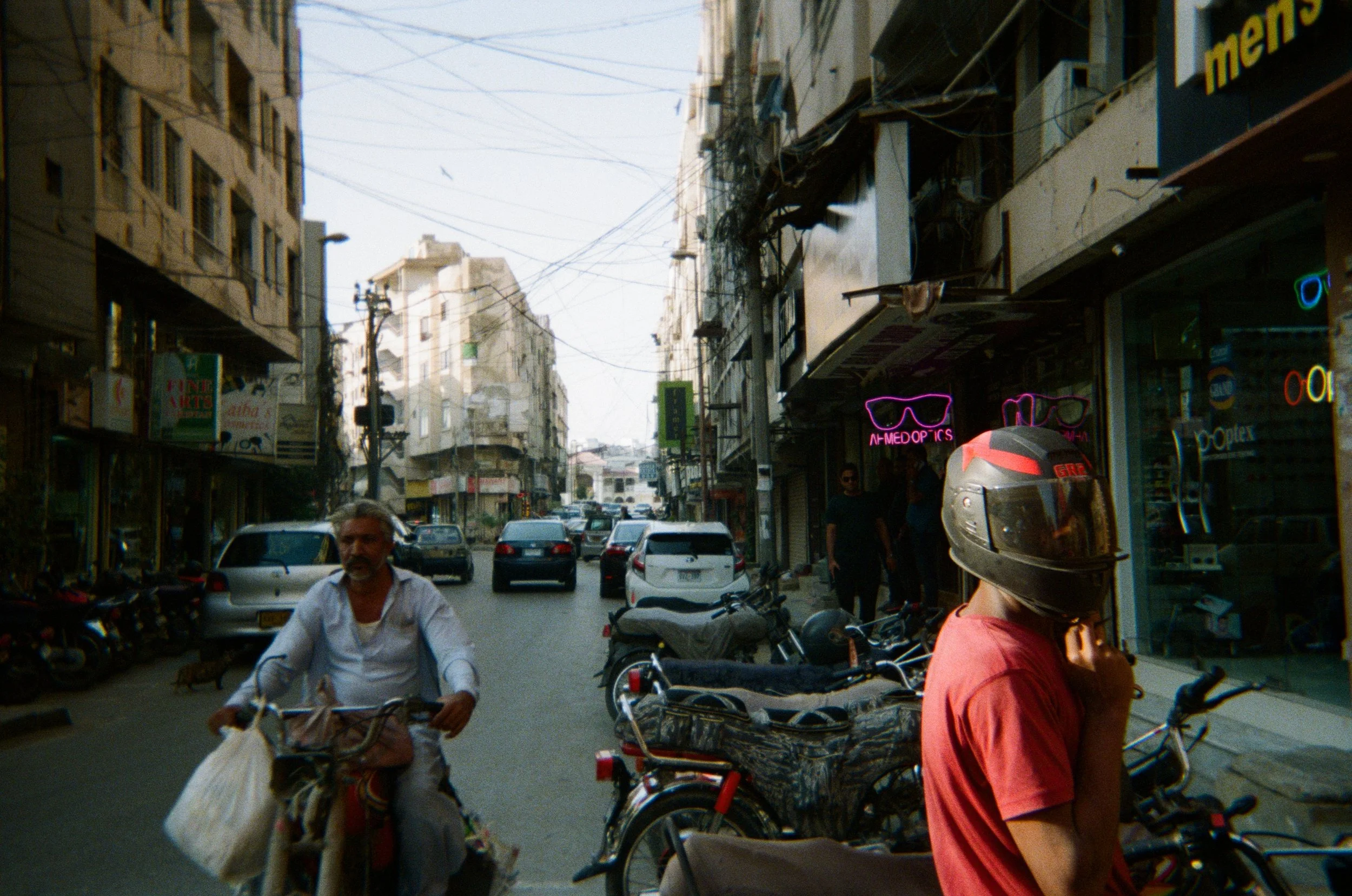 A busy street in Pakistan lined with shops and parked motorcycles, with a neon eyeglasses sign and a person in a helmet in the foreground — photographed by the STUDIO PURANI founder on a sourcing trip