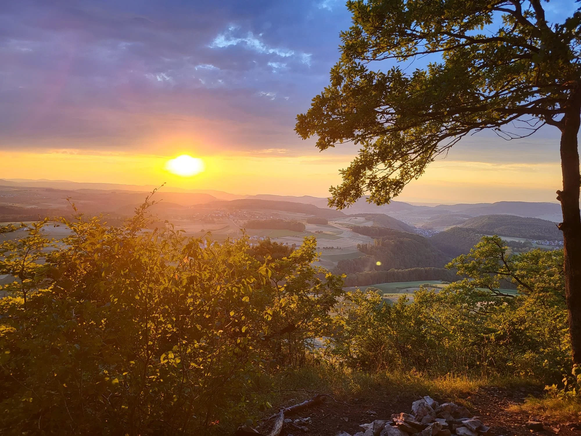 Ein malerischer Sonnenuntergang über einer hügeligen Landschaft mit Bäumen und Feldern.