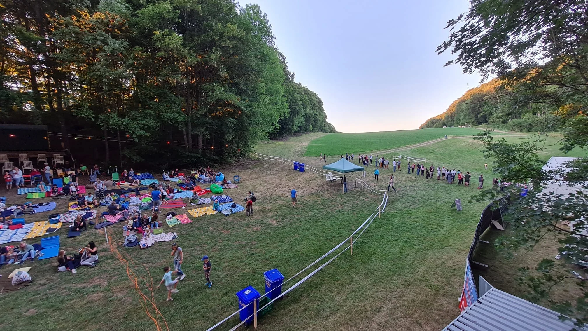 Ein Sommerfest im Freien mit Menschen, die sich auf der Wiese entspannen, auf Decken sitzen, und in einer Warteschlange stehen. Es gibt Tische, einen Pavillon, einen Bereich mit Sand und eine umgebende Baumlinie.