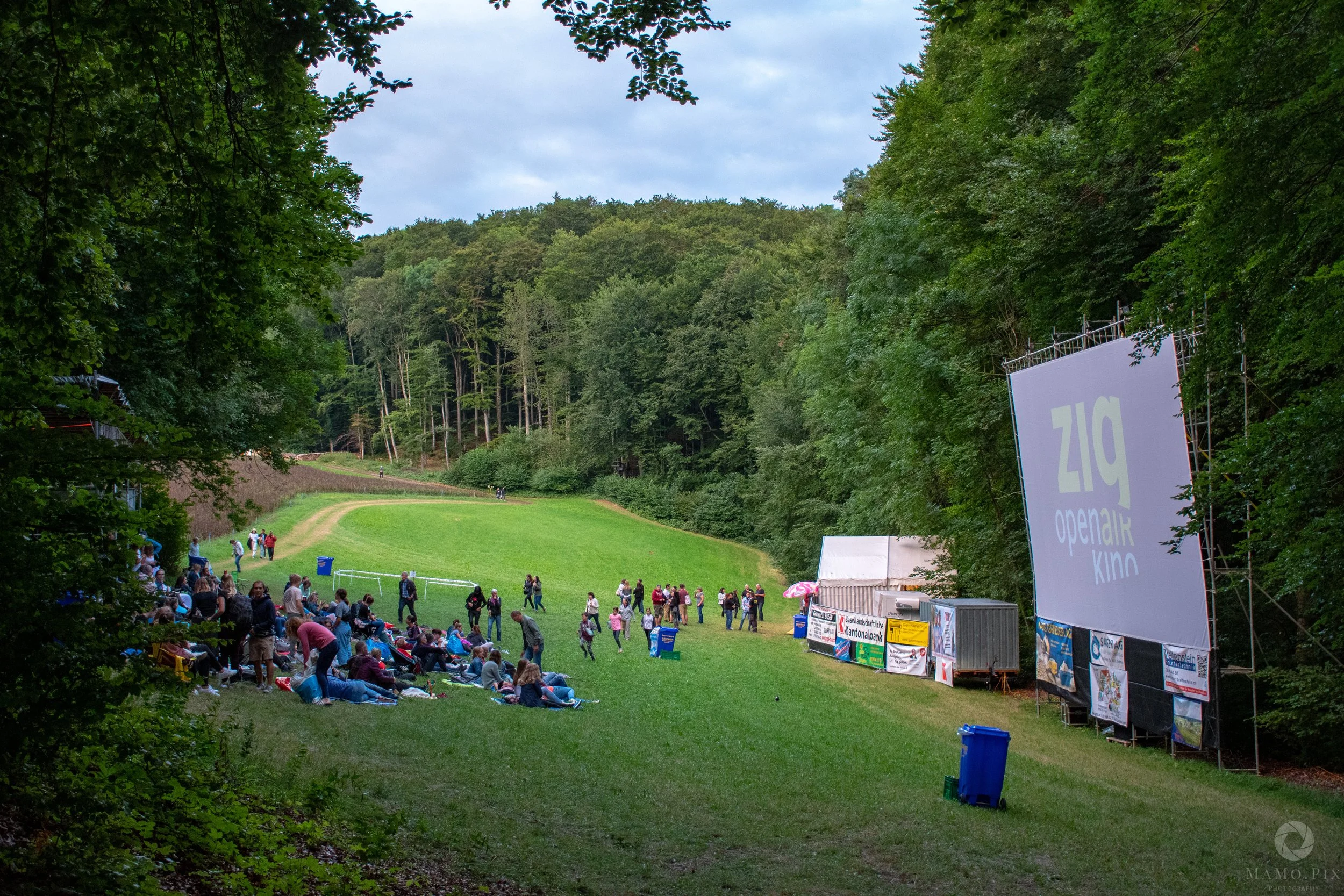 Gruppe von Menschen sitzt und steht im Freien vor einer großen Kinoleinwand, die in einem bewaldeten Park aufgestellt ist, für ein Open-Air-Kino-Event bei Tageslicht.