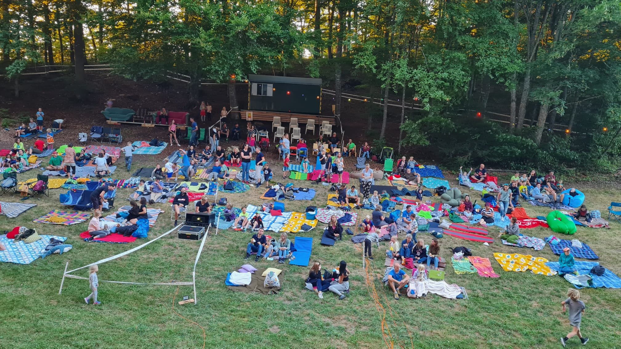 Viele Menschen sitzen auf farbigen Decken im Freien, umgeben von Bäumen, mit einem kleinen Konzertbühne und Sitzgelegenheiten im Hintergrund.