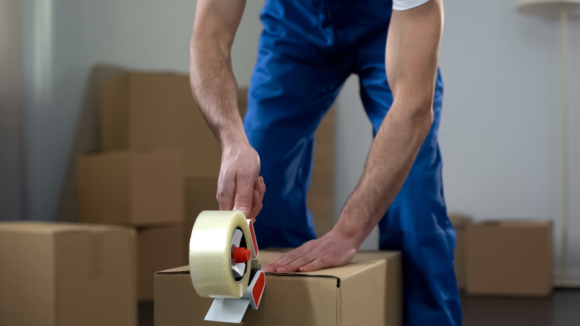 A person wearing blue pants and a white shirt taping cardboard boxes with packing tape in a storage or warehouse setting.