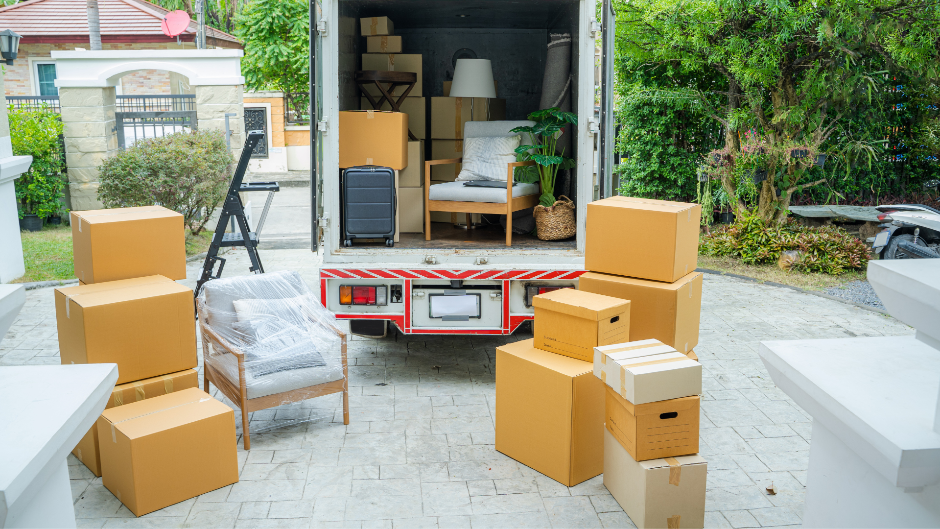 Moving truck with packed boxes, furniture, and household items outside in a driveway during daylight.