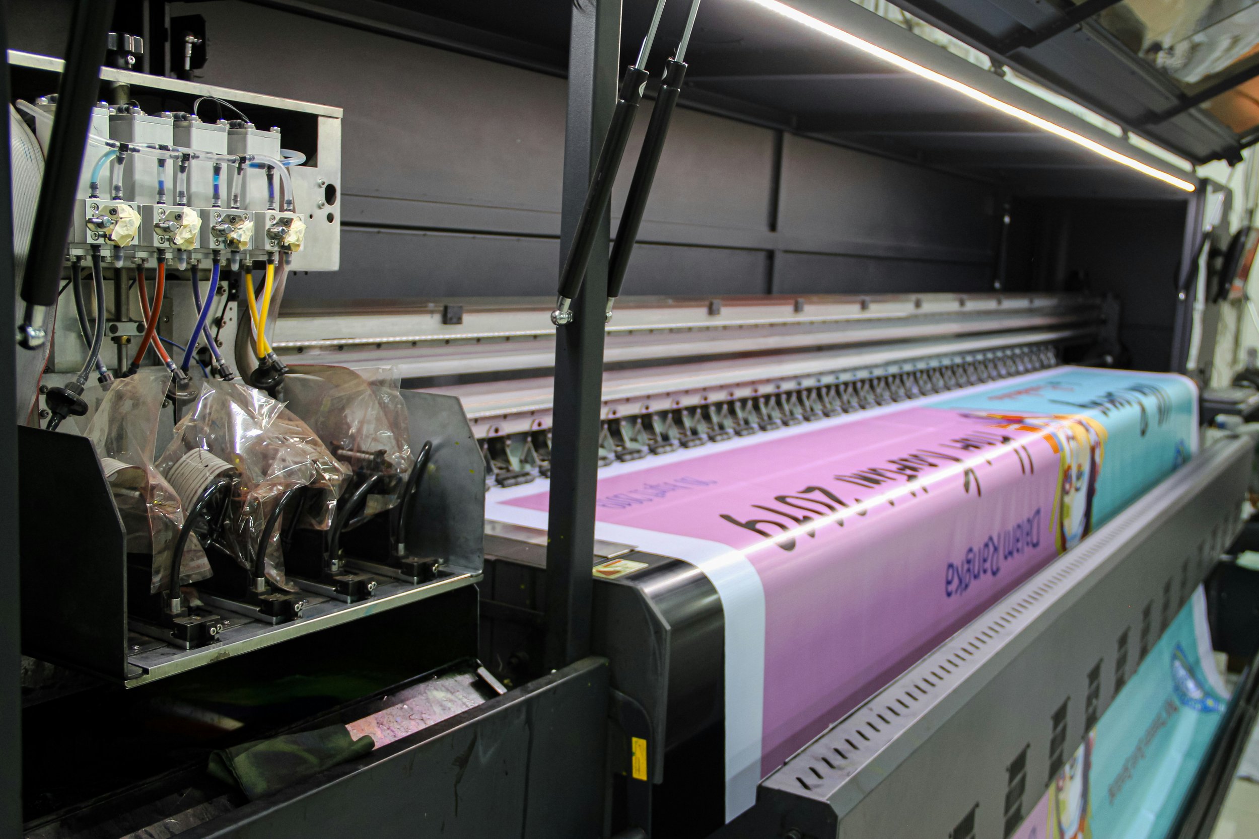 Close-up of a large industrial printer printing a colorful poster or banner with pink, light blue, and orange sections, with various wires and mechanical components visible on the left side.