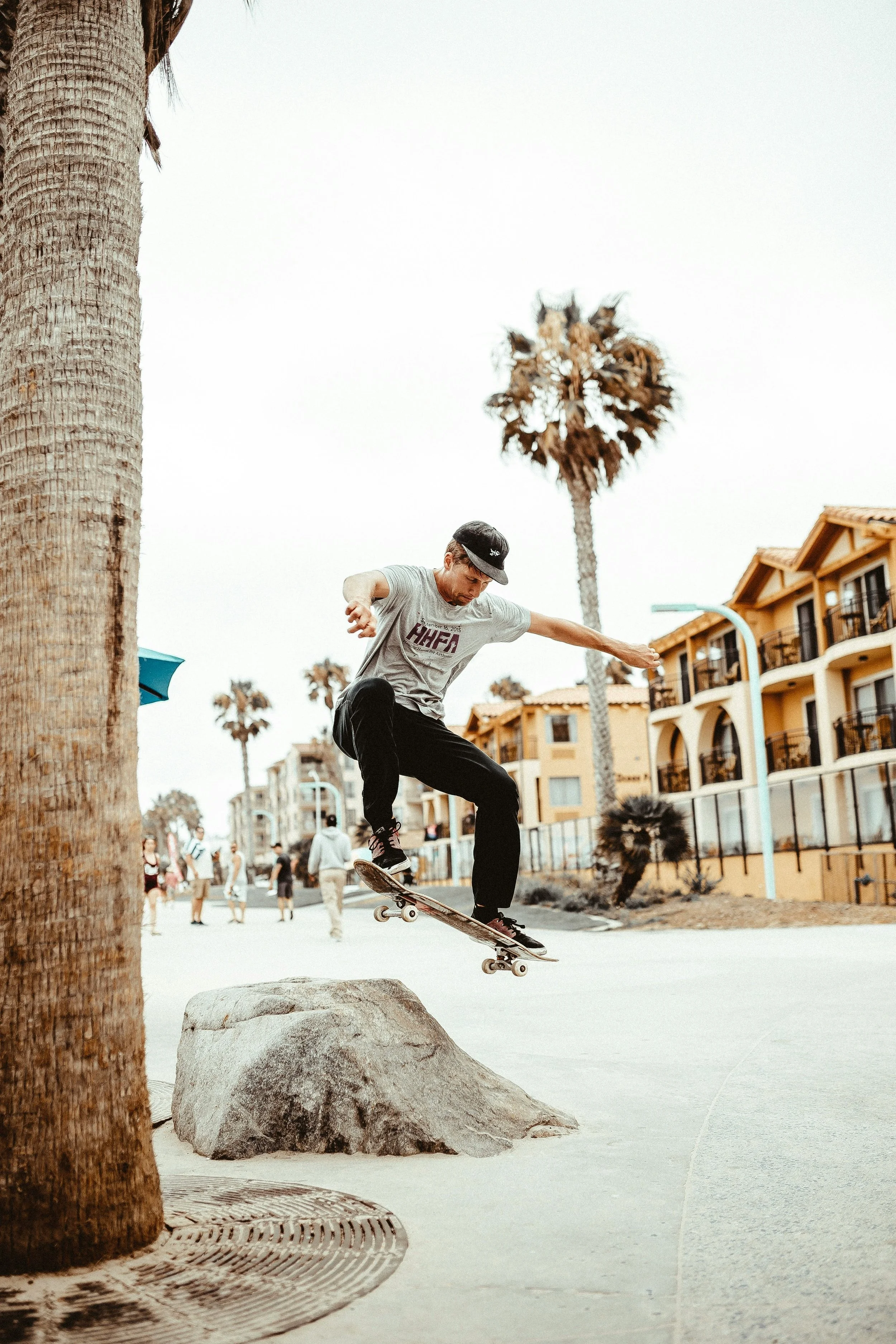 Skateboarder performing a trick on a rock at a beachside promenade with palm trees and apartment buildings in the background.