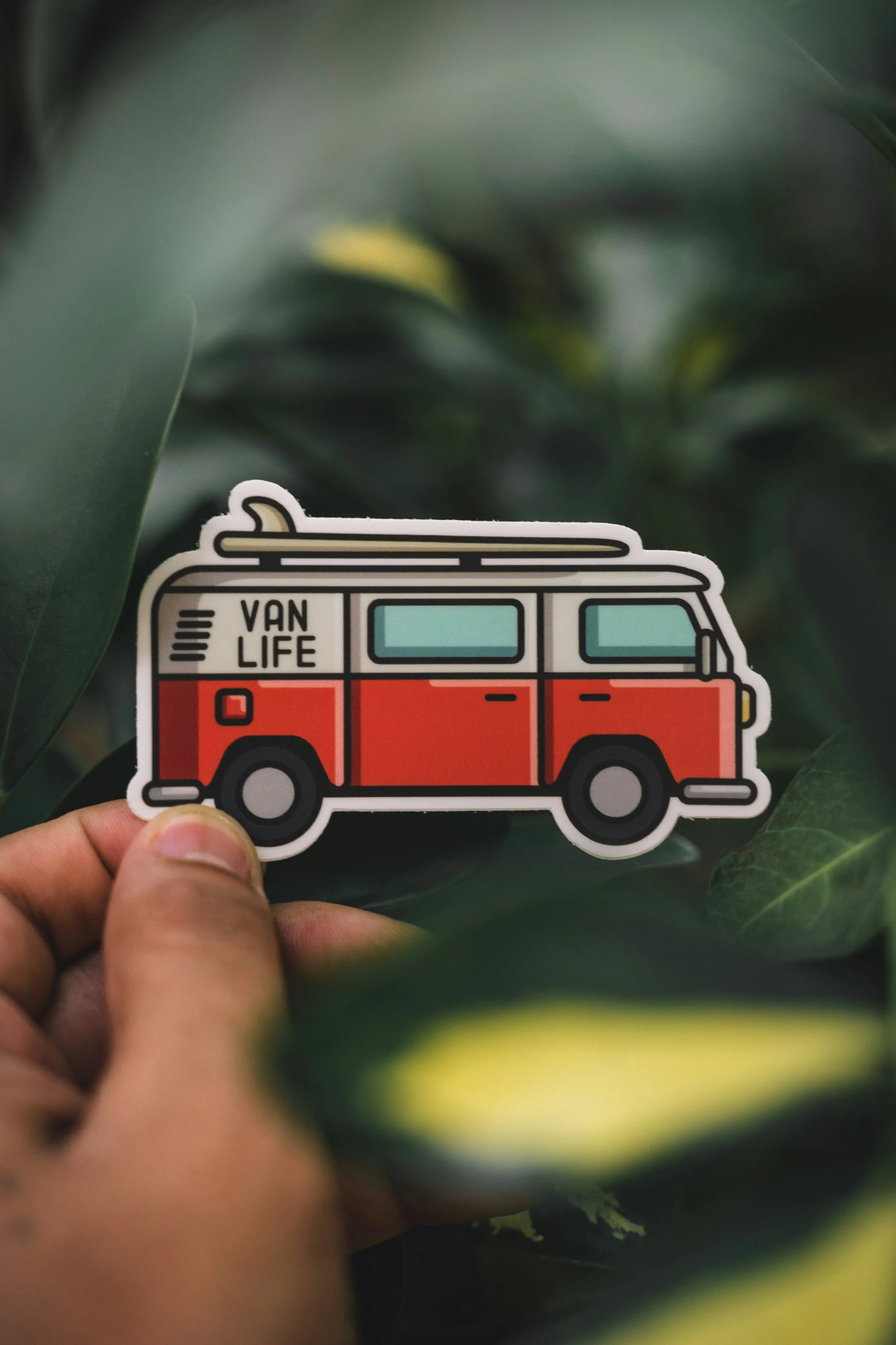 A hand holding a sticker of a red and cream vintage van with a surfboard on top, with the words 'VAN LIFE' written on the side, against a background of green leaves.