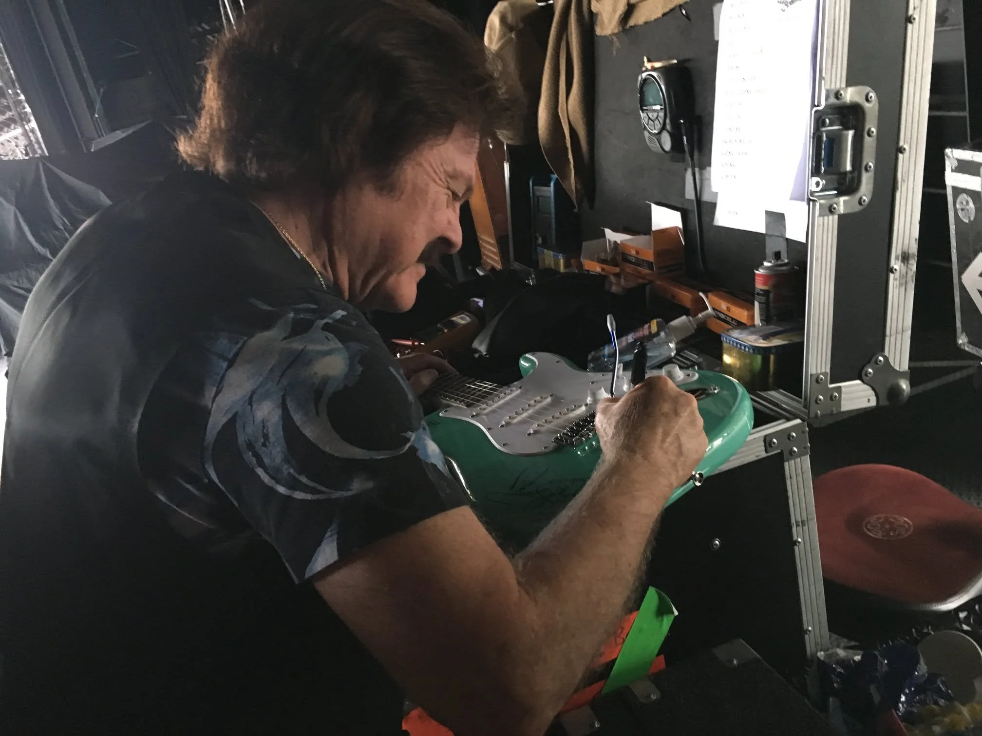 A man working on an electric guitar in a workshop or studio setting. The workspace has various tools, papers, and electronic equipment.