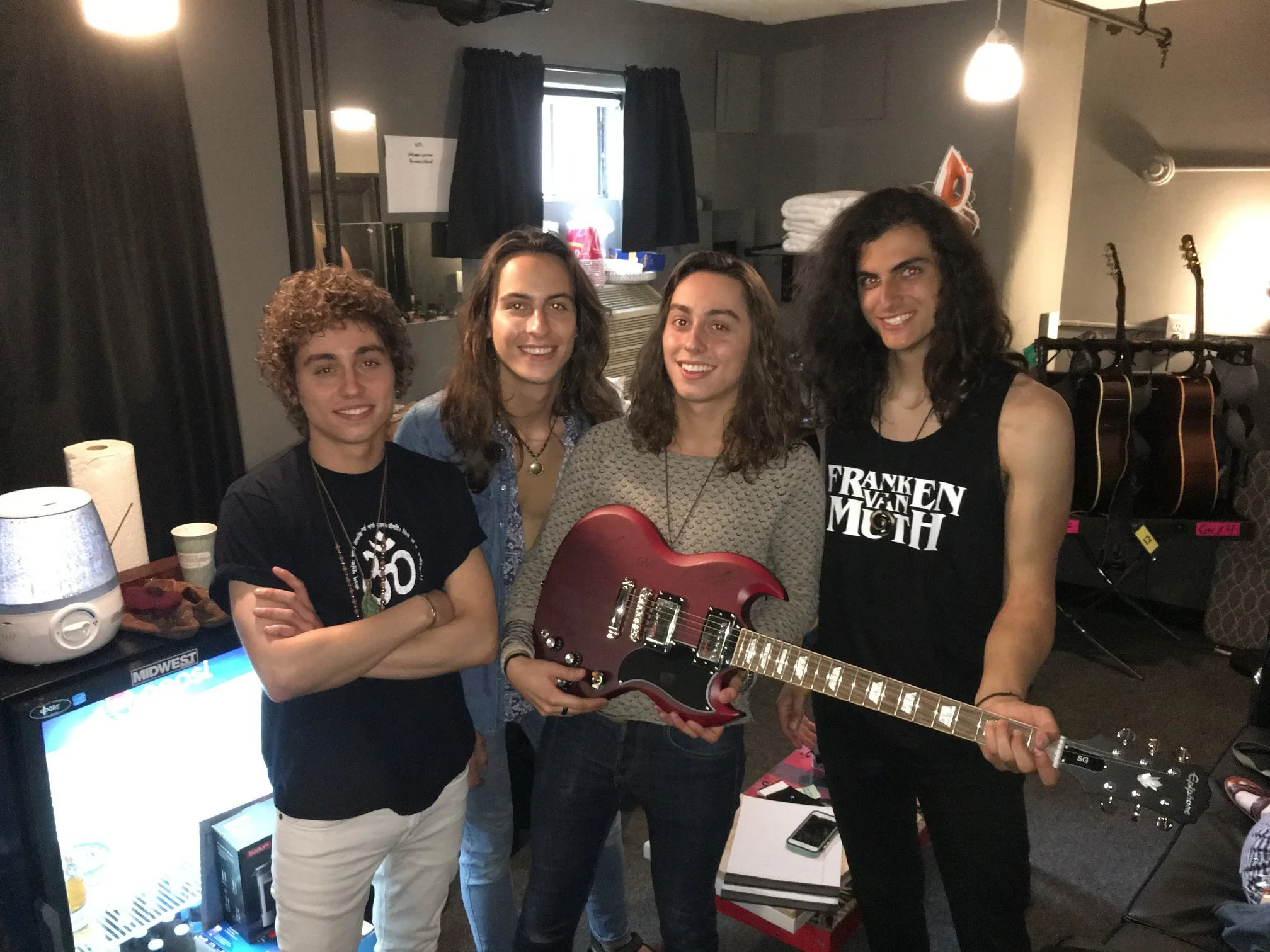 Four people standing together in a room, one of whom is holding a pink electric guitar. The background features a window with dark curtains, a stack of white towels, a desk with electronics, and multiple guitars on stands.