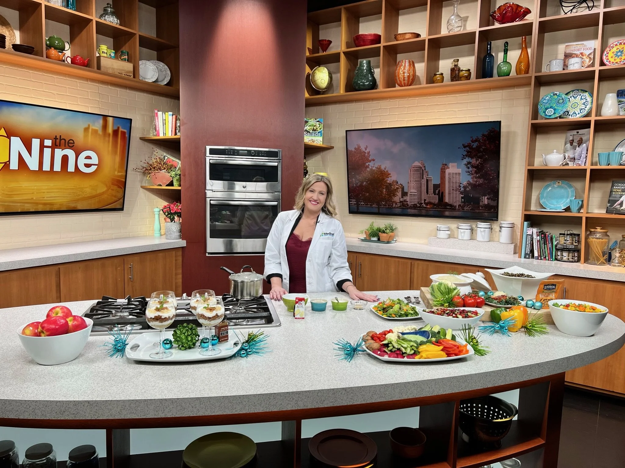 A woman in a white coat smiling in a kitchen set with baked goods, salads, and vegetables on a large counter decorated with blue ornaments and greenery, with shelves filled with colorful pottery and books in the background.