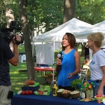 Woman in blue dress being interviewed by a cameraman at an outdoor event with a table of food and drinks