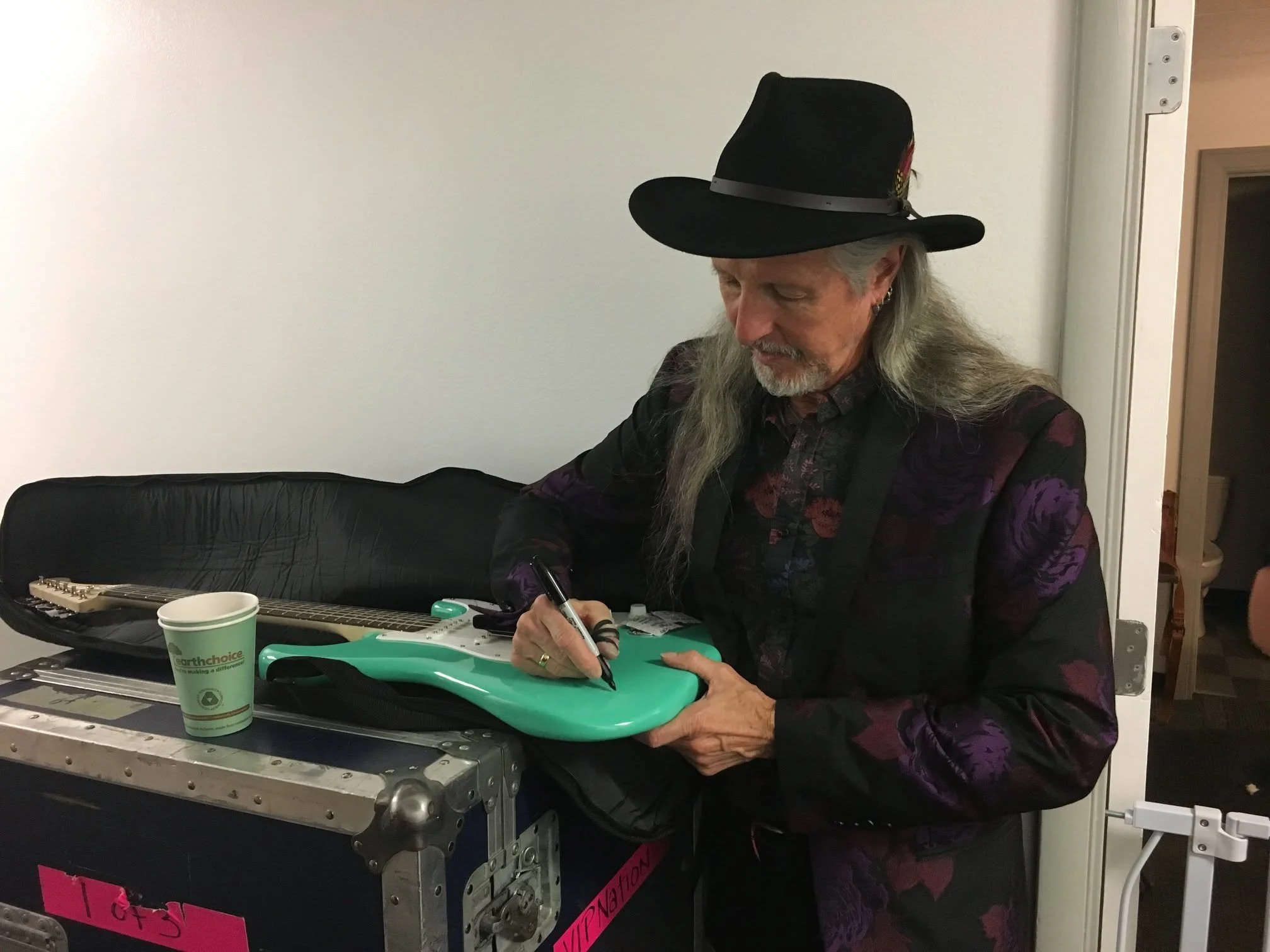 A man with long gray hair and a gray beard wearing a black wide-brimmed hat, dark floral shirt, and earrings, is sitting at a table signing an object. On the table are a black guitar case, a paper cup, and some tools. The background shows a white wall and a doorway.
