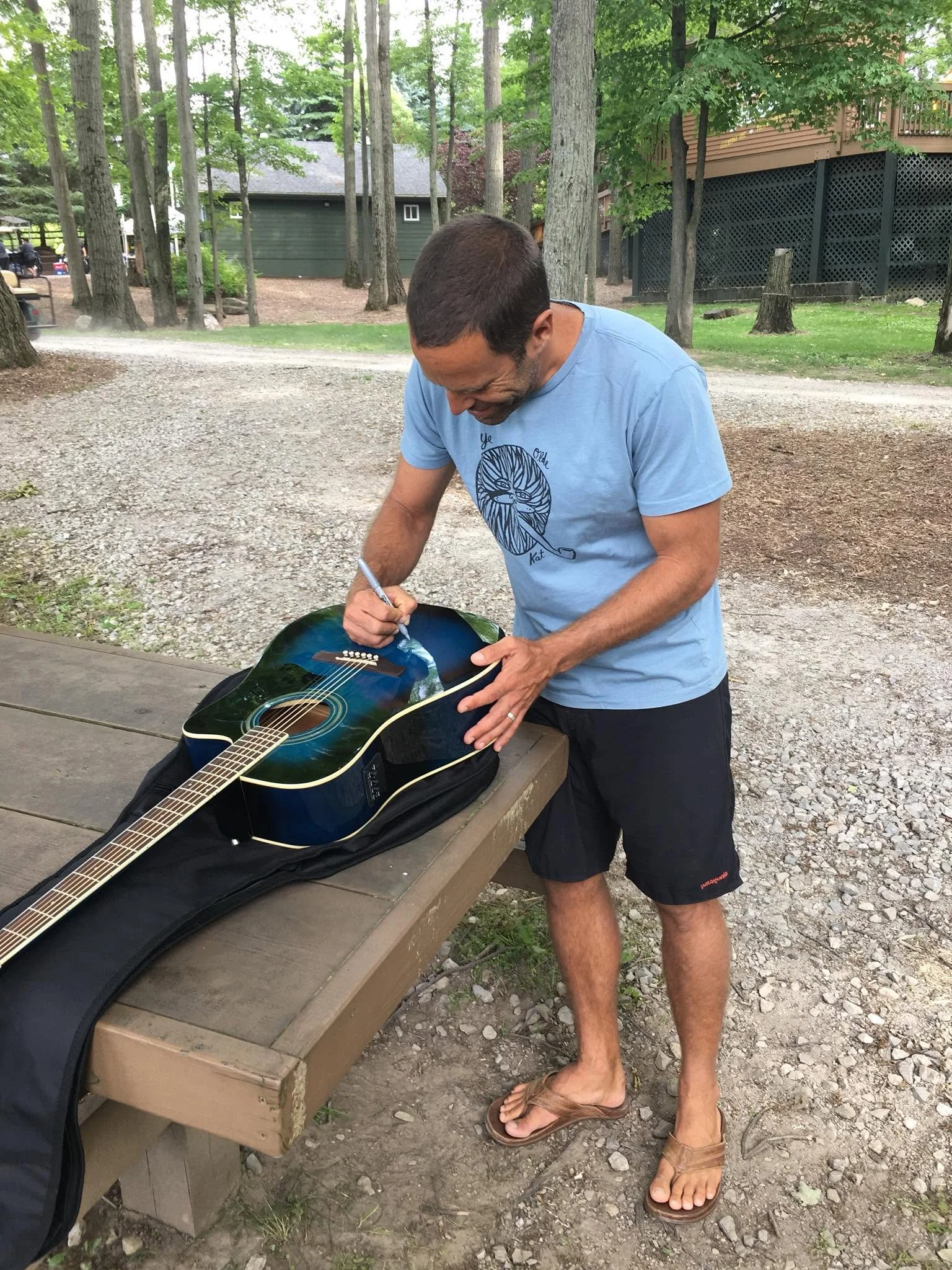A man with short dark hair is signing a colorful acoustic guitar that is resting on a wooden bench. He is wearing a light blue T-shirt with a graphic design, black shorts, and brown sandals. The setting appears to be outdoors in a wooded area with trees, a gravel path, and a green building in the background.