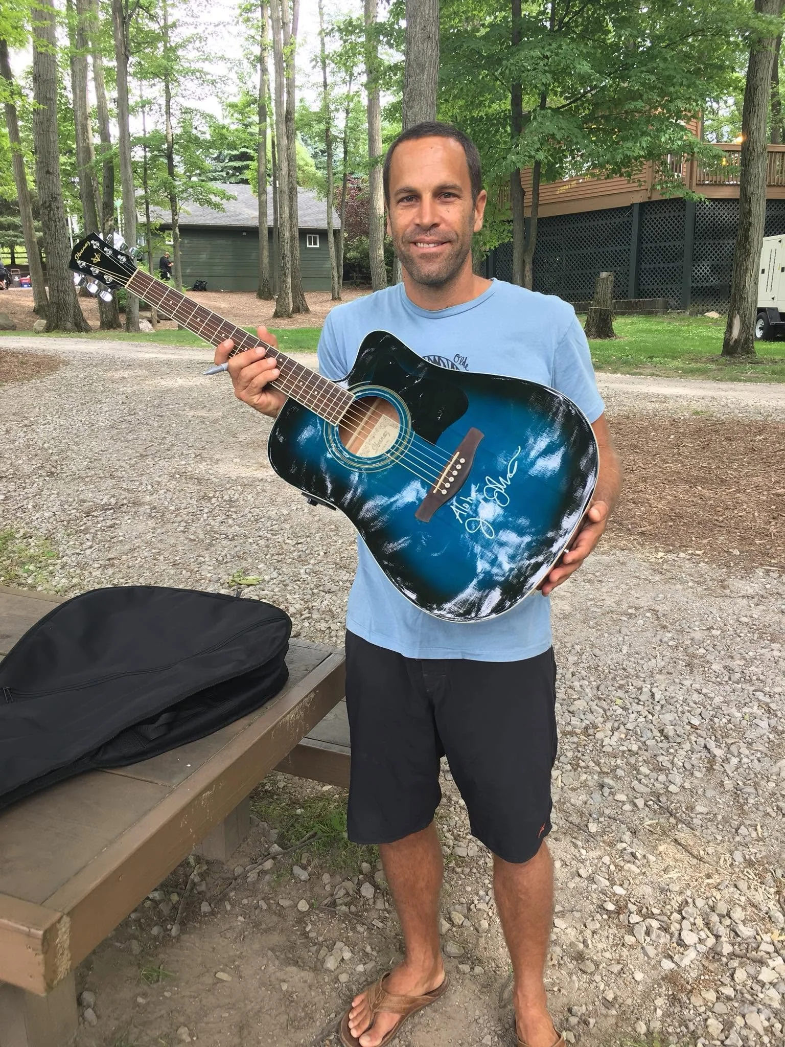 A man stands outdoors in a wooded area holding an autographed blue and black acoustic guitar, smiling at the camera. There is a bench with a black bag on it nearby and trees in the background.