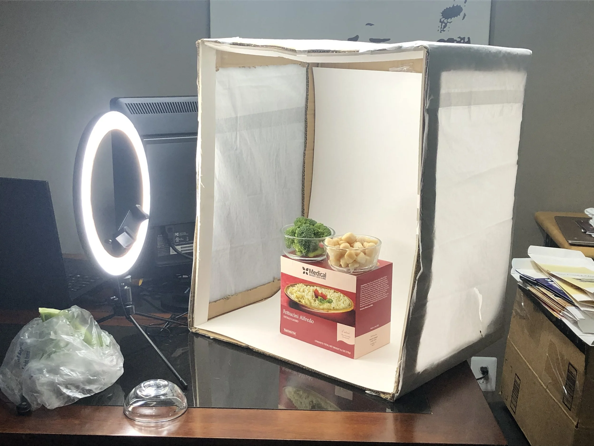 A cardboard box with a cardboard interior, placed on a black table, containing a small bowl of garlic cloves and a small bowl of broccoli, with a ring light reflecting off the table.