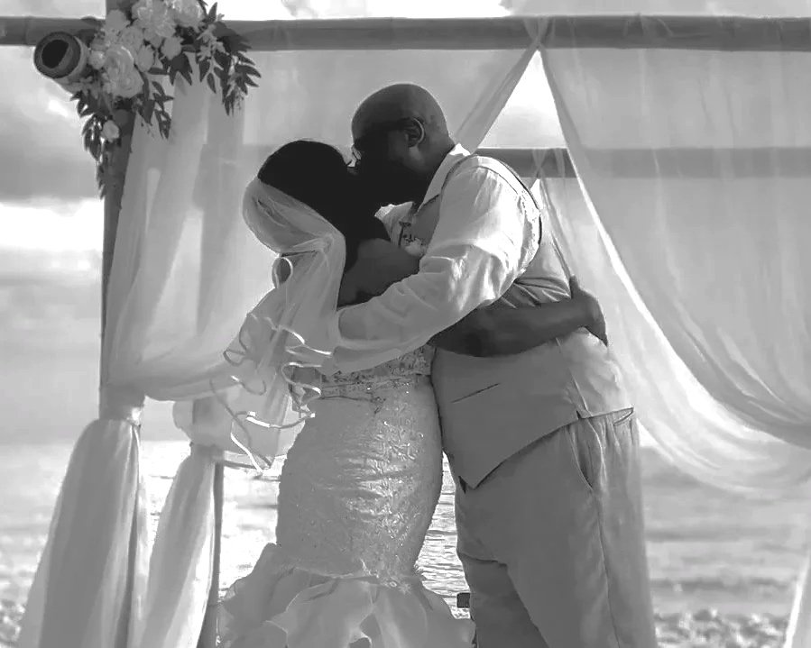A black-and-white photo of a man in a suit and a bride in a wedding dress, standing close together outdoors.