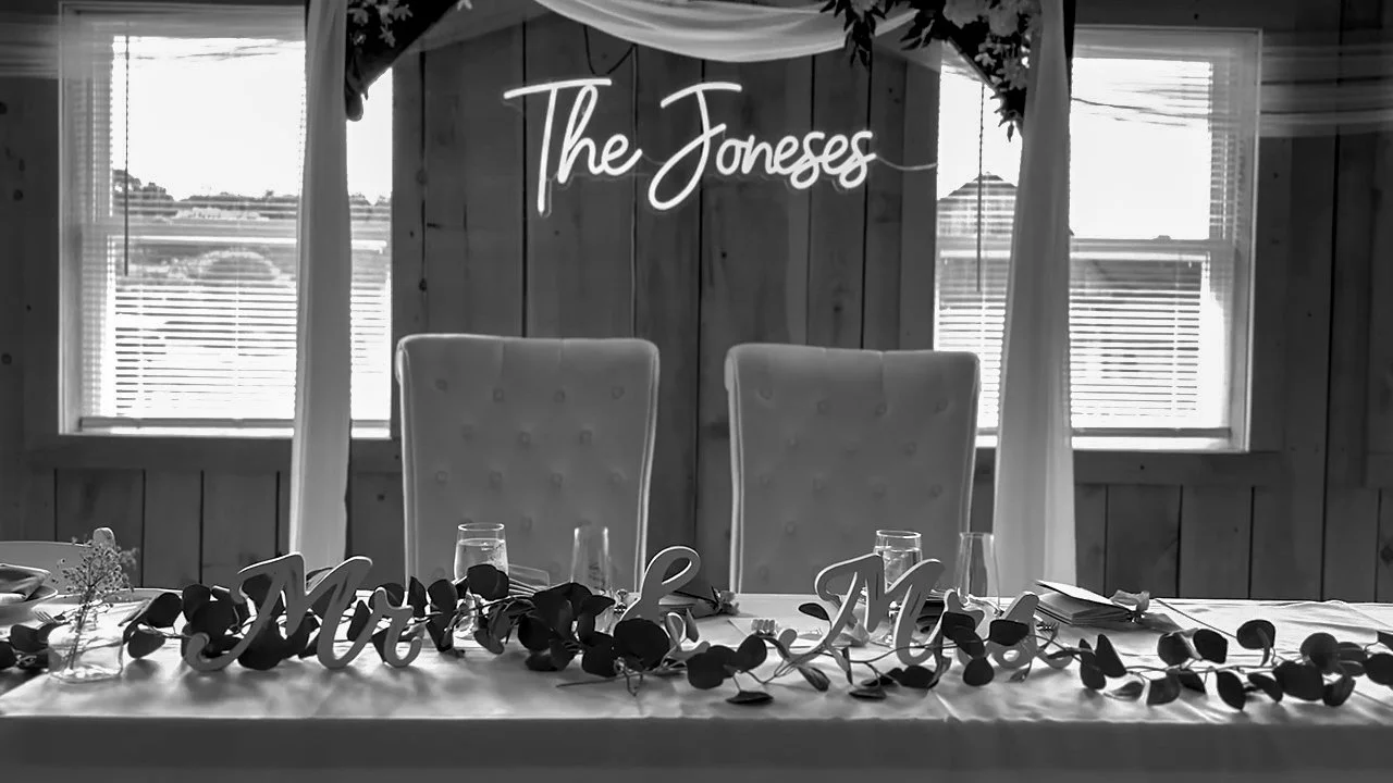 A rustic indoor setting with a wooden wall background, featuring a sign that says 'The Joneses' and sitting at a table with chairs. There are windows letting in natural light and some decorations on the table.
