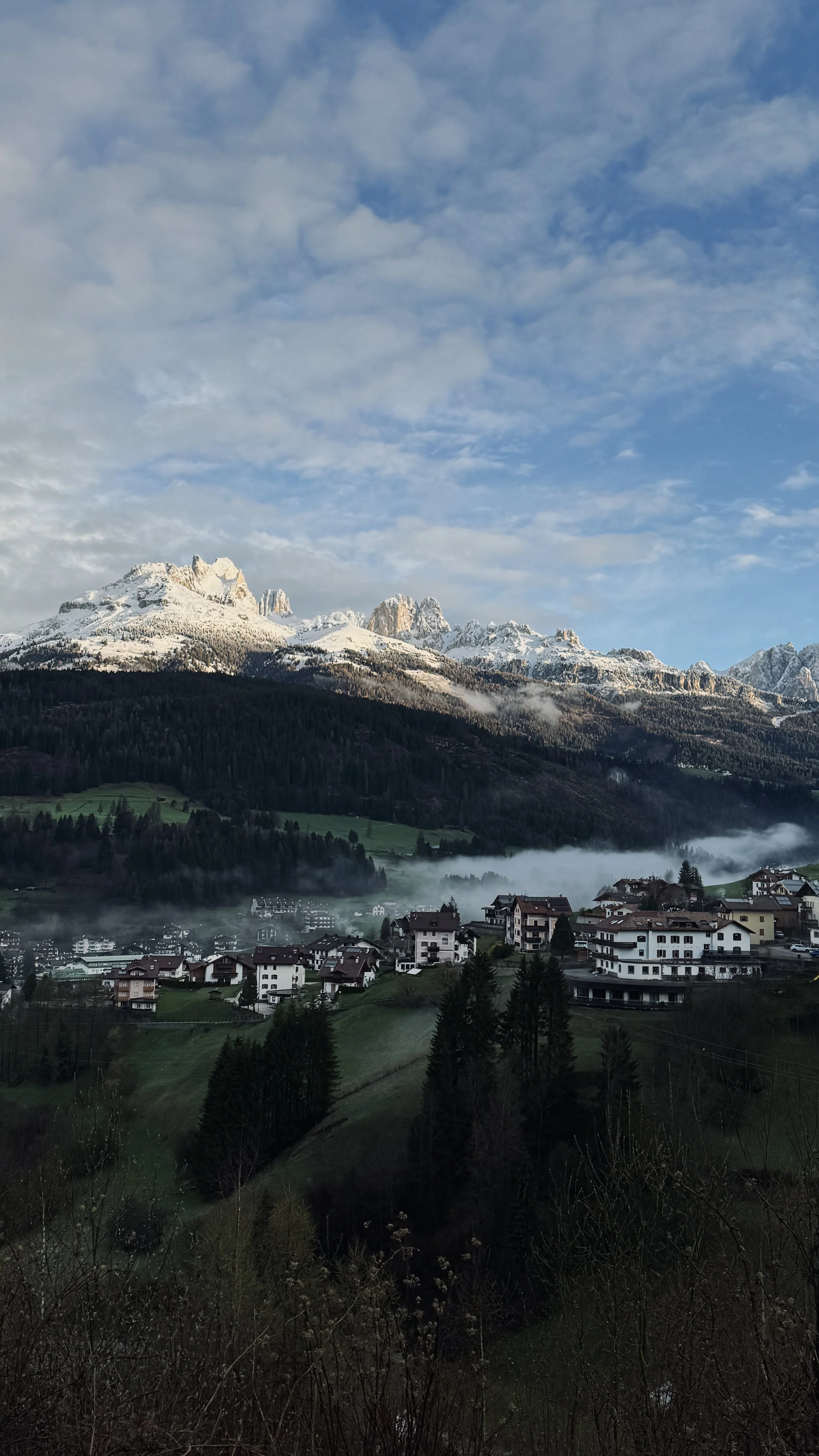 Vista de un pueblo en un valle rodeado de montañas con nieve, árboles y casas, con cielo parcialmente nublado