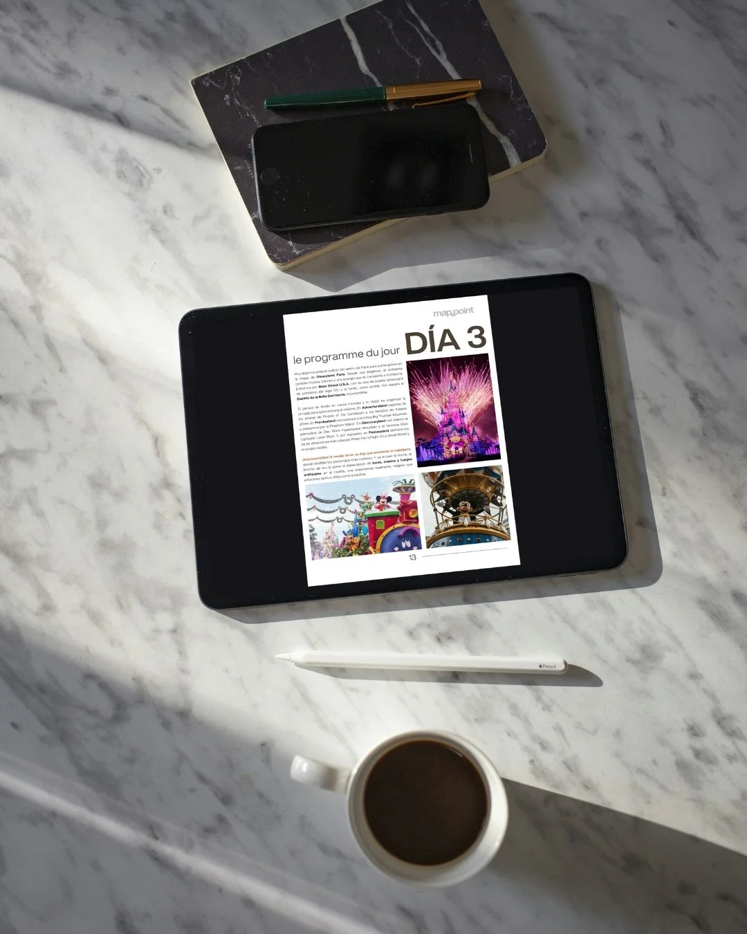 Tableta con una revista digital en la pantalla mostrando un programa del día 3 con imágenes de parques temáticos, una taza con café, un bolígrafo, un teléfono móvil y un cuaderno sobre una mesa de mármol.