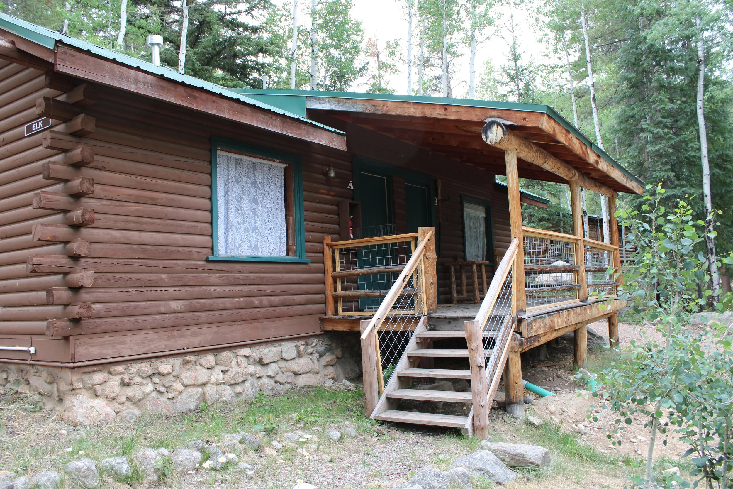 A rustic wooden cabin with a small front porch, surrounded by trees and greenery.