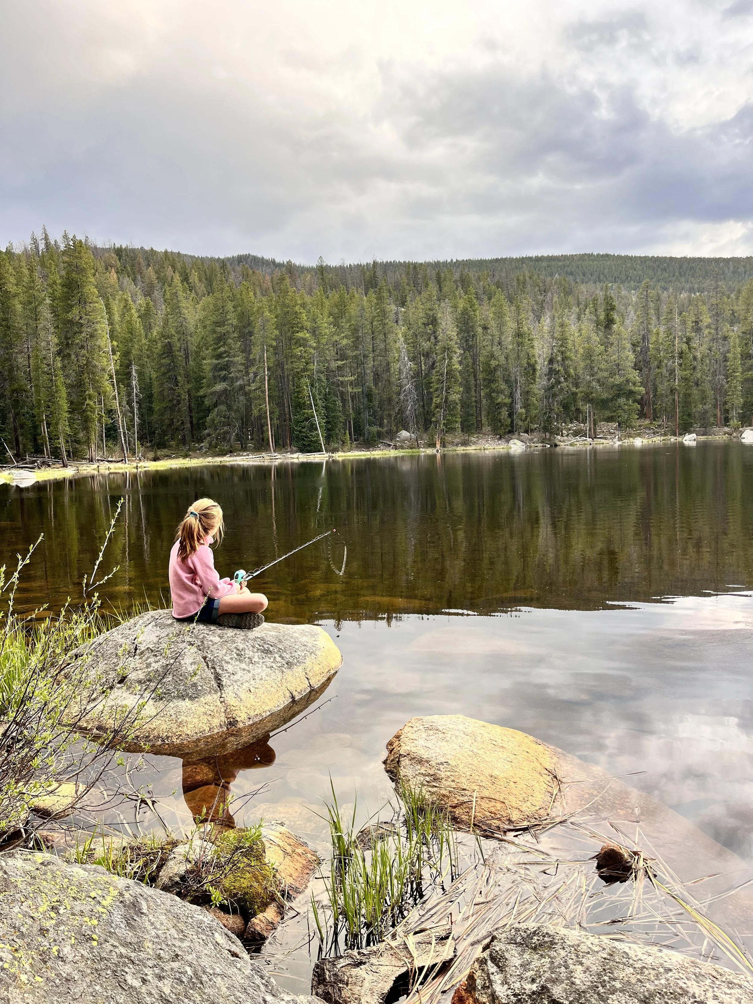 A young girl with blonde hair sitting on a large rock by a lake, fishing with a small fishing rod. The lake is surrounded by a dense forest of tall green trees under a partly cloudy sky.