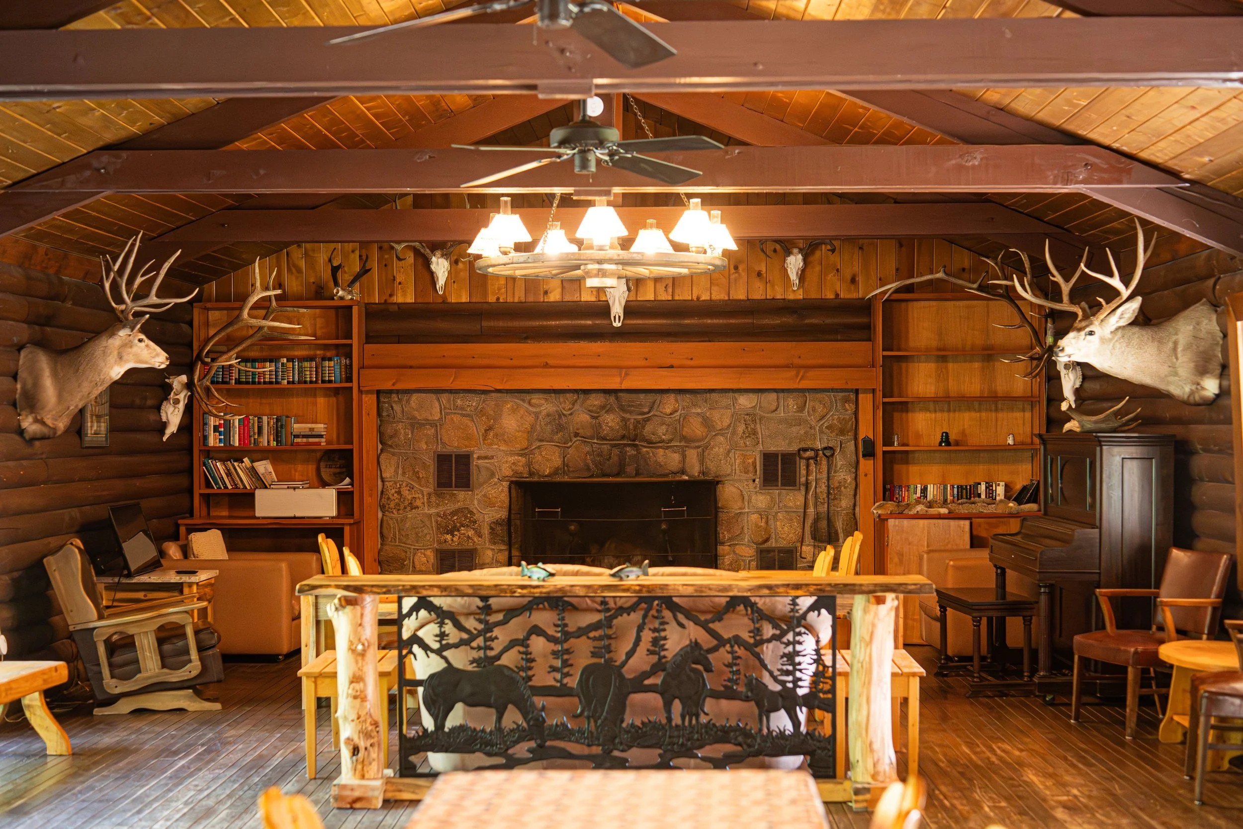 Interior of a rustic cabin with a stone fireplace, wooden ceiling, and walls decorated with mounted elk heads and skulls, featuring a chandelier and a piano.