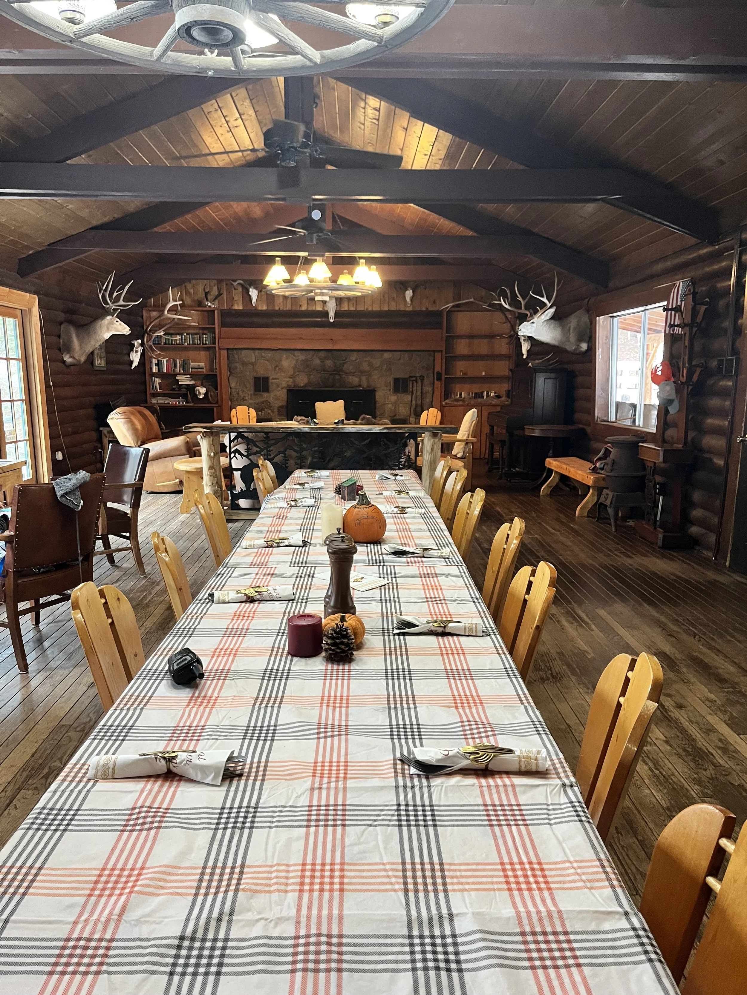 A long dining table with a plaid tablecloth set with napkins, utensils, and centerpieces including a pumpkin, pinecone, candles, and a salt shaker. The room features wood-paneled walls, a ceiling with exposed beams, mounted deer heads, and large windows. There's a stone fireplace at the far end with bookshelves on either side, and the space has a rustic cabin decor.