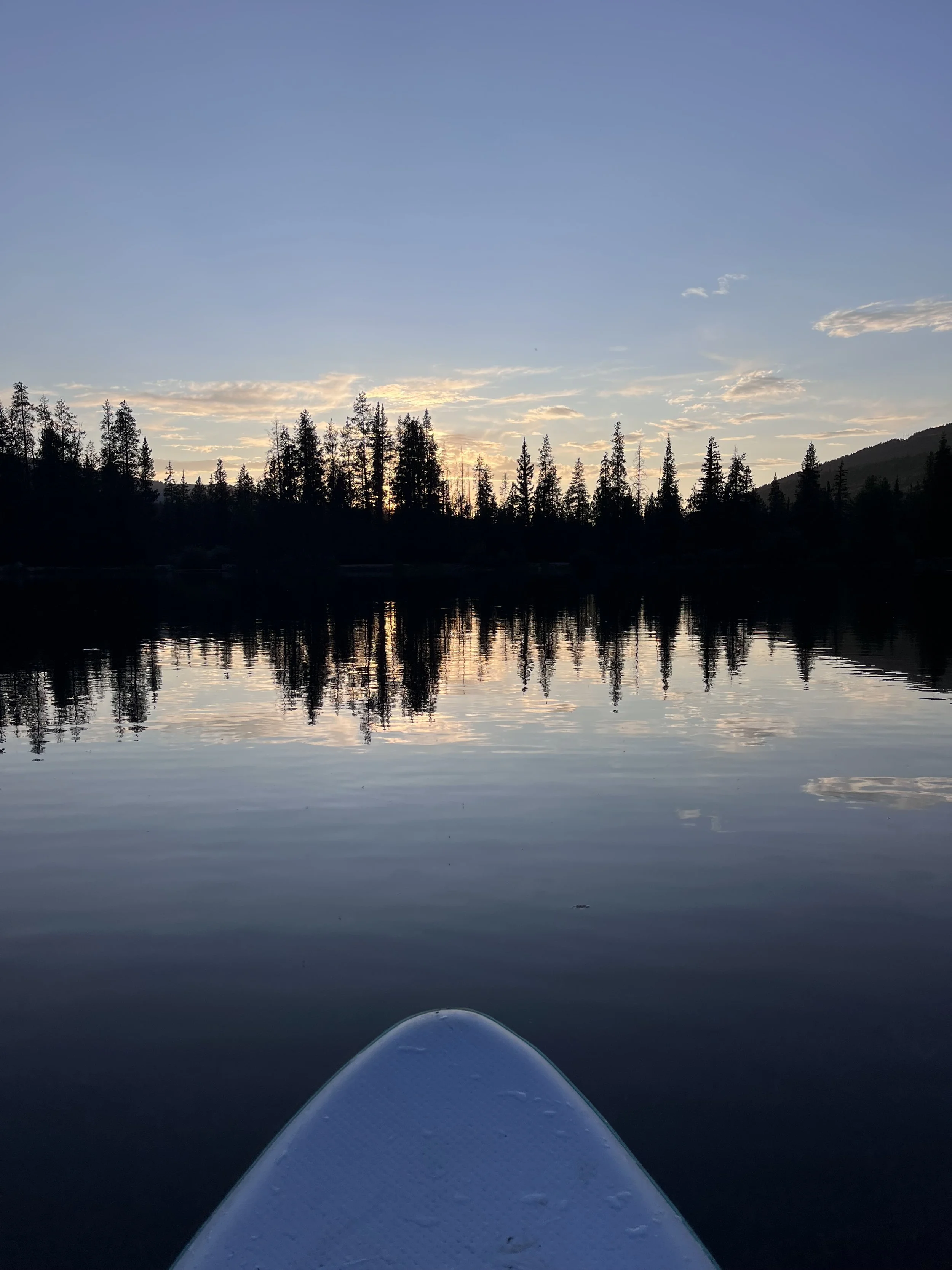 View from a paddleboard on a calm lake at sunset, with the sun setting behind a forested shoreline and mountain in the background, reflecting on the water.