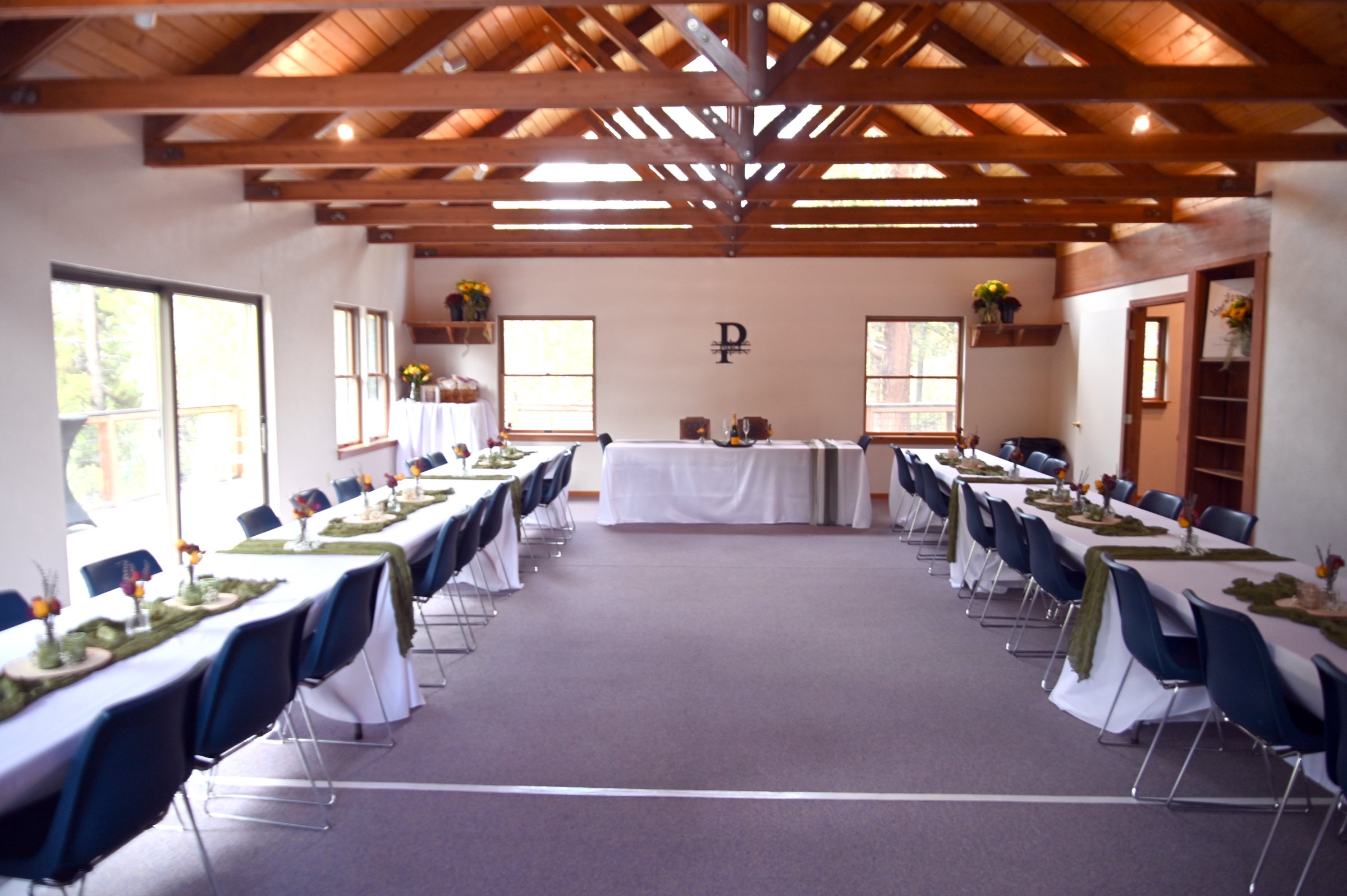 A wedding reception setup with long tables covered in white tablecloths, decorated with small vases of flowers and green table runners, arranged in a U-shape in a rustic hall with wooden beams on the ceiling and daylight coming through windows.