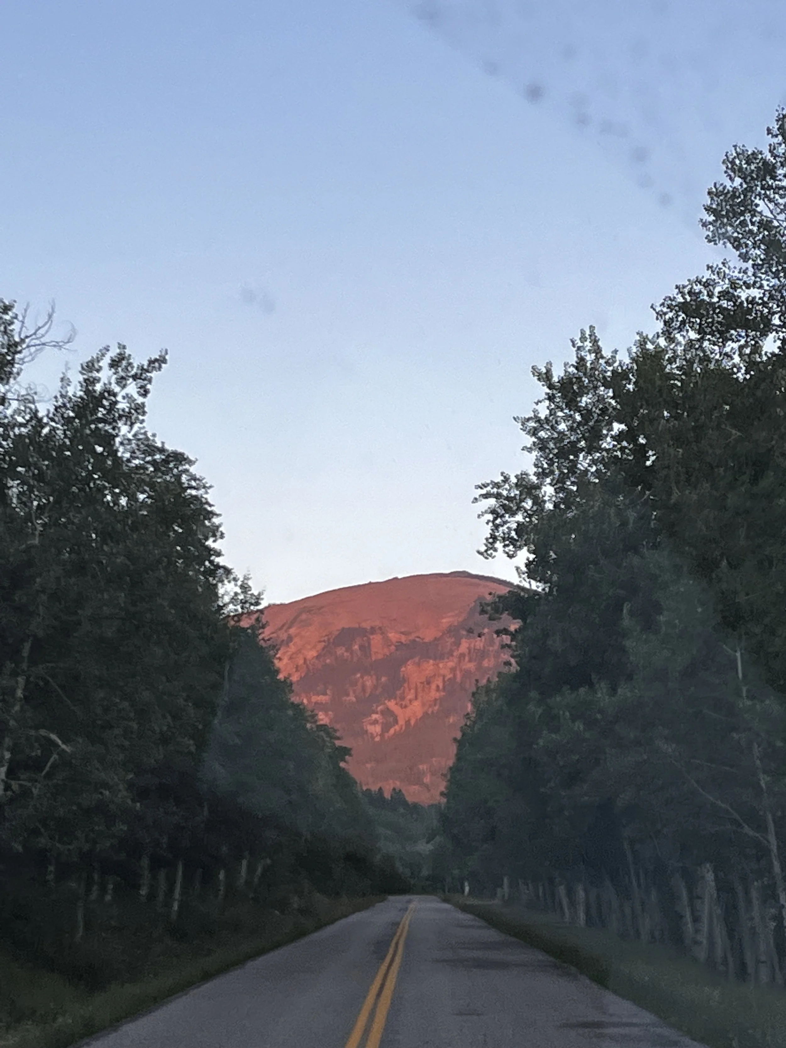 A two-lane road lined with trees on both sides, leading toward a mountain illuminated by the setting or rising sun, with a clear blue sky above.