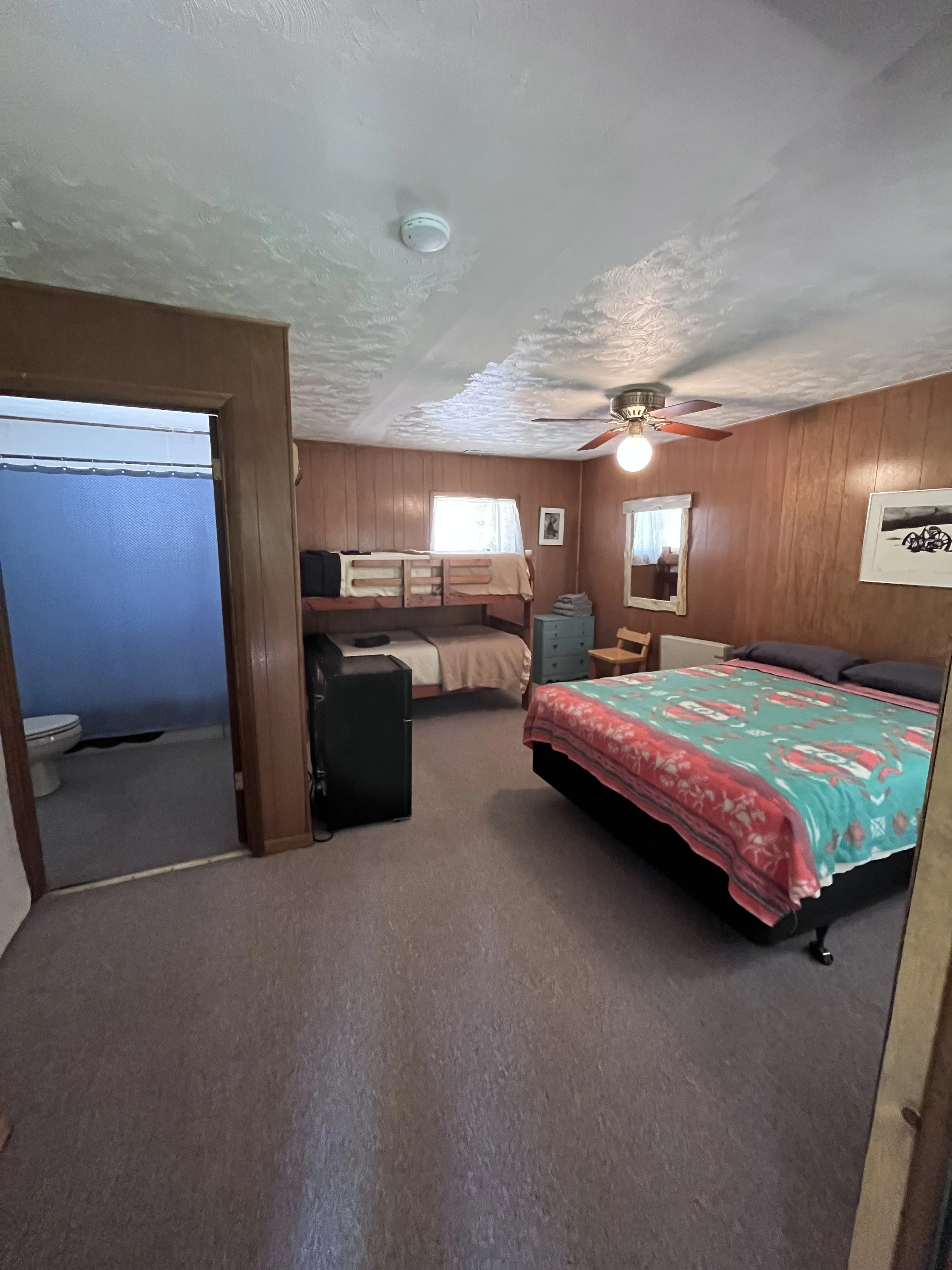 Wood-paneled bedroom with a large bed covered with a colorful blanket, a ceiling fan with a light, a bunk bed in the corner, small window, and a bathroom entrance with a blue shower curtain.