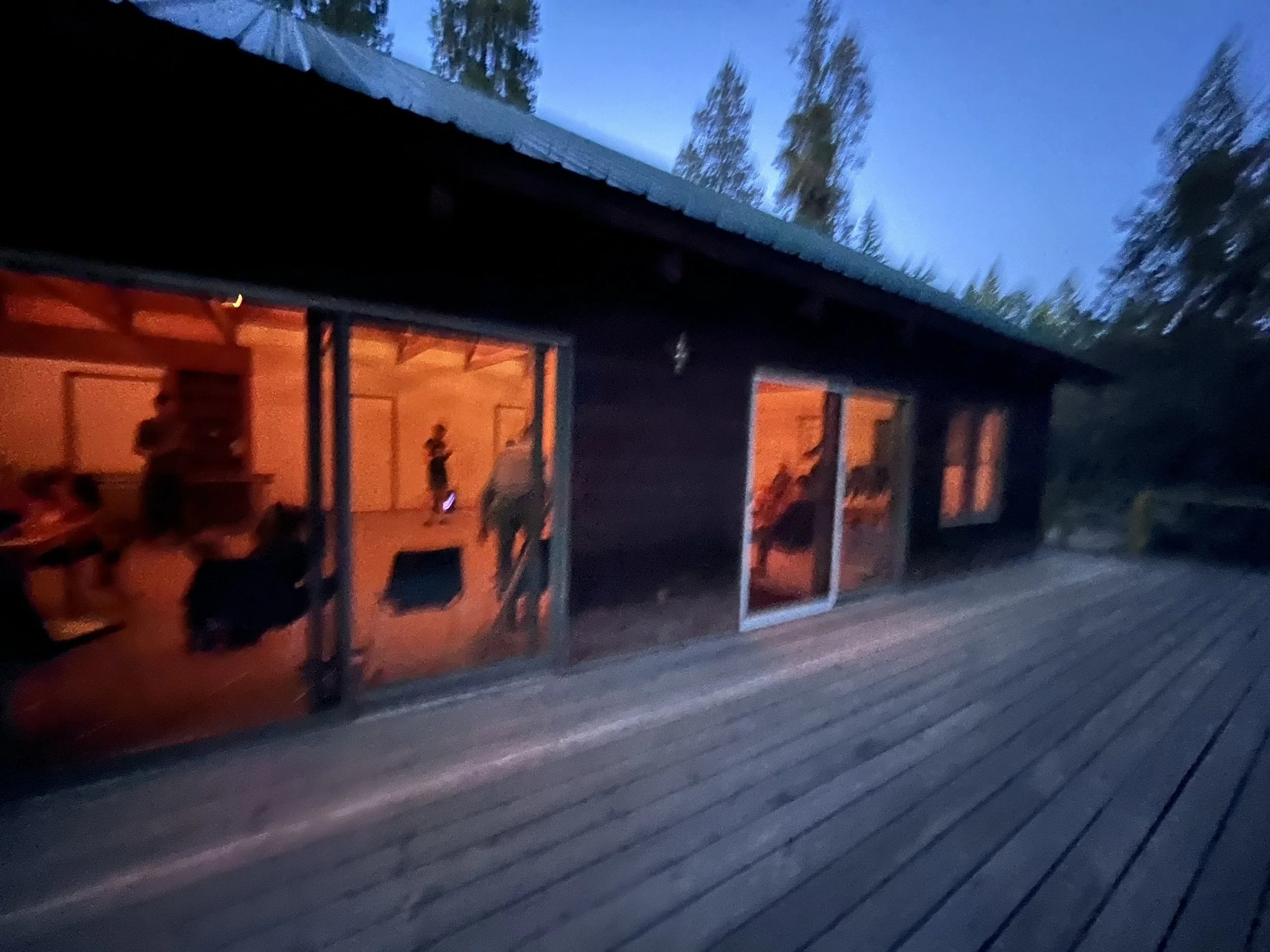 People inside a cabin with large glass sliding doors, illuminated with warm orange lighting, surrounded by trees during dusk or early evening.