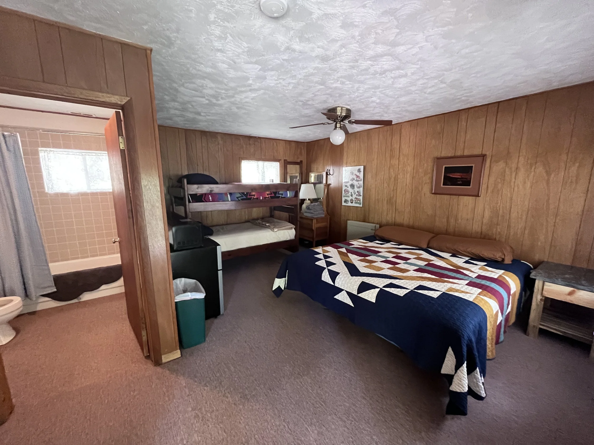 A bedroom with wood-paneled walls, a large bed with a quilted blanket and two pillows, a ceiling fan, a bunk bed, a small dresser with stacked towels, and a framed picture on the wall.
