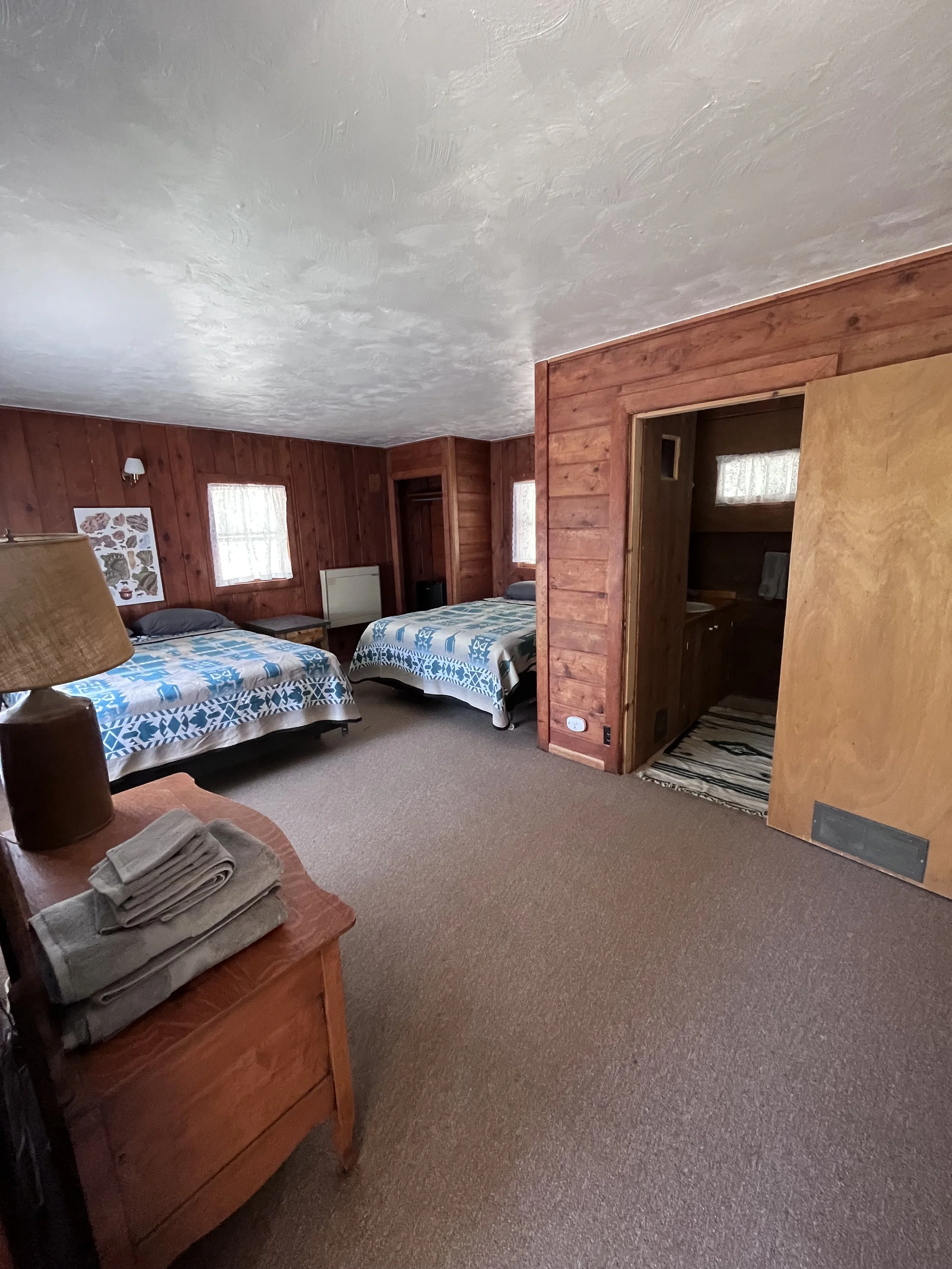 A cozy bedroom with two beds with blue and white patterned covers, wood-paneled walls, two small windows with lace curtains, a nightstand, a painting on the wall, a small bathroom doorway, and folded towels on a dresser.