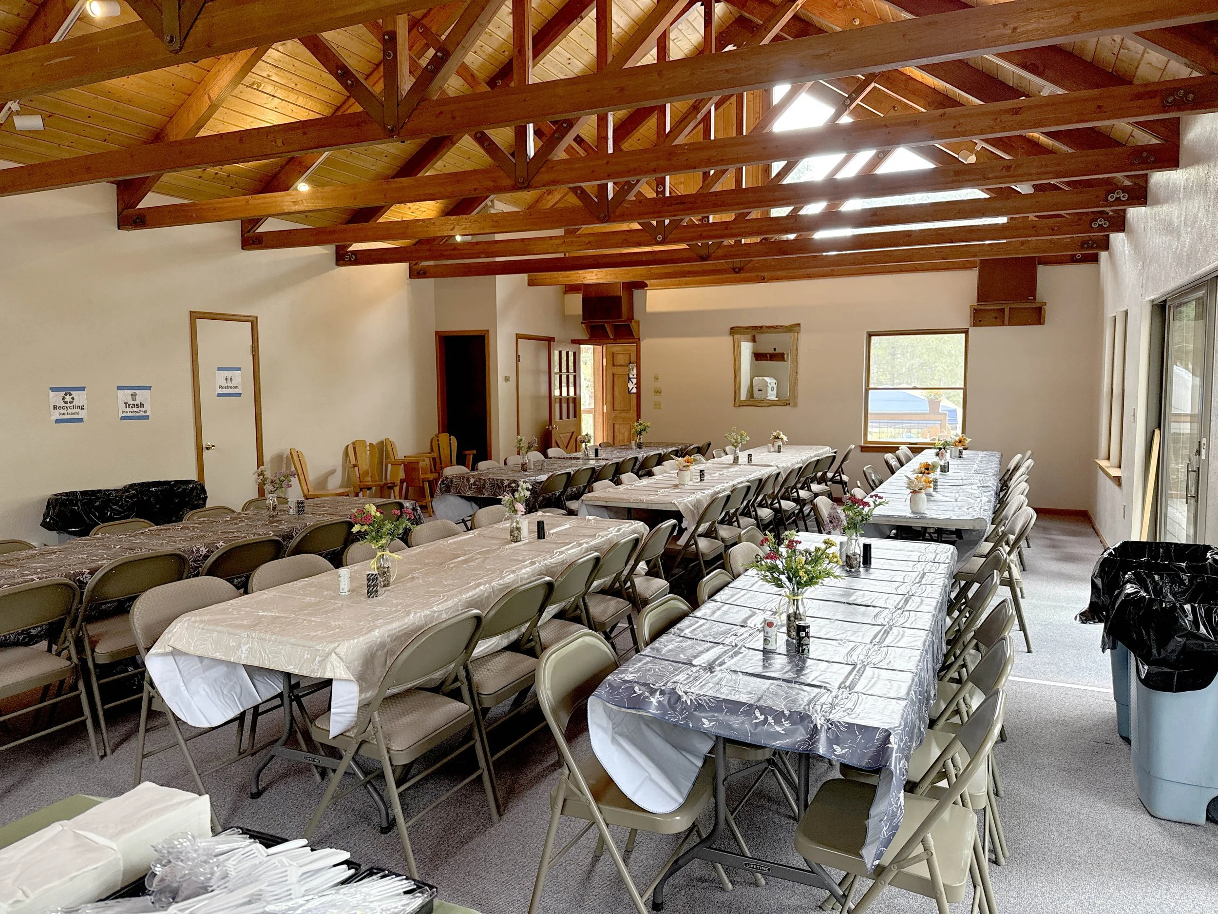 A large dining hall with several long tables covered in plastic and floral arrangements in small vases. The hall has a high wooden ceiling with exposed beams and windows along the walls. There are trash and recycling bins at the edges of the room.