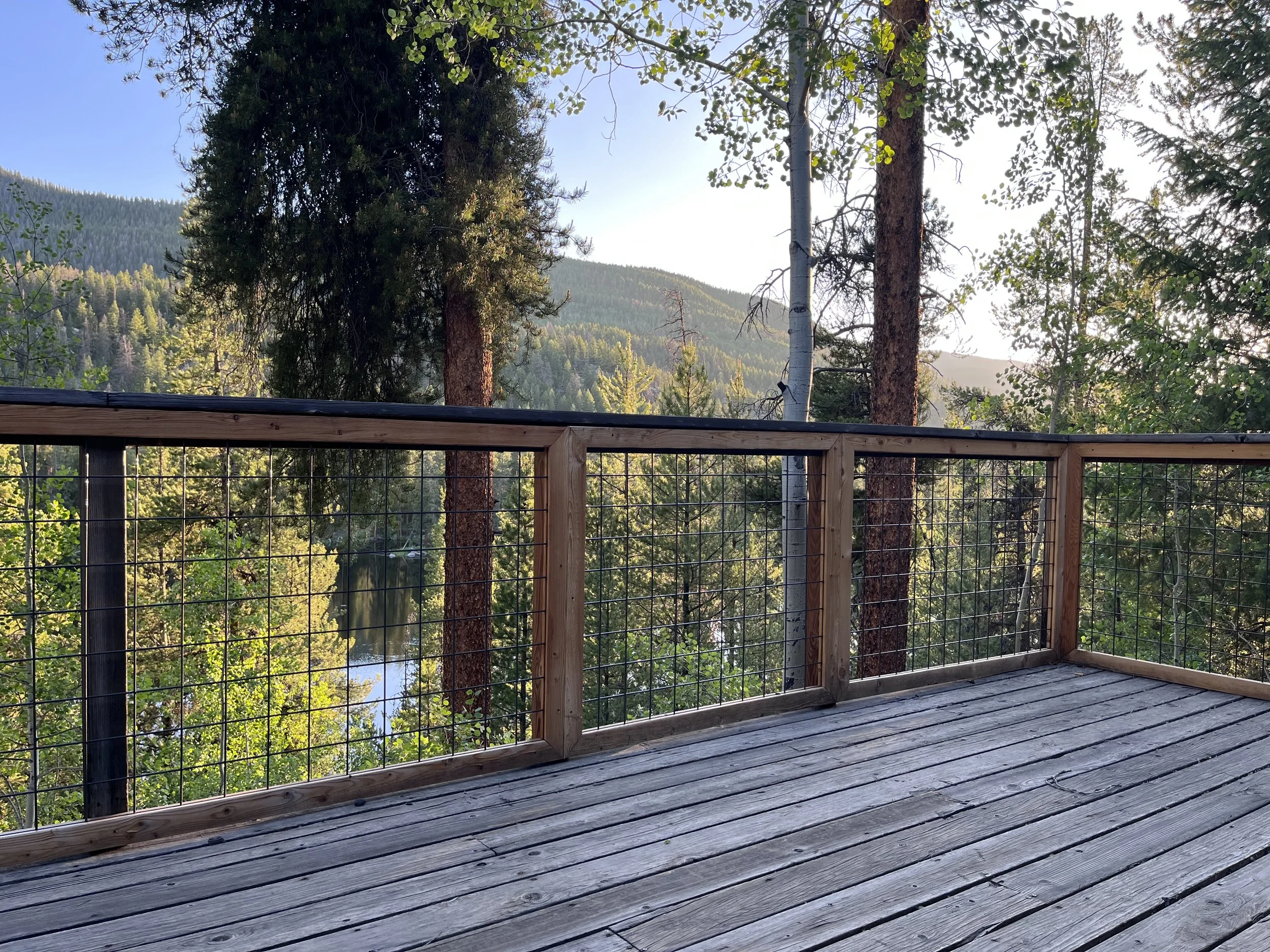 A wooden deck with a metal wire railing overlooking a forested landscape with trees, mountains, and a river.
