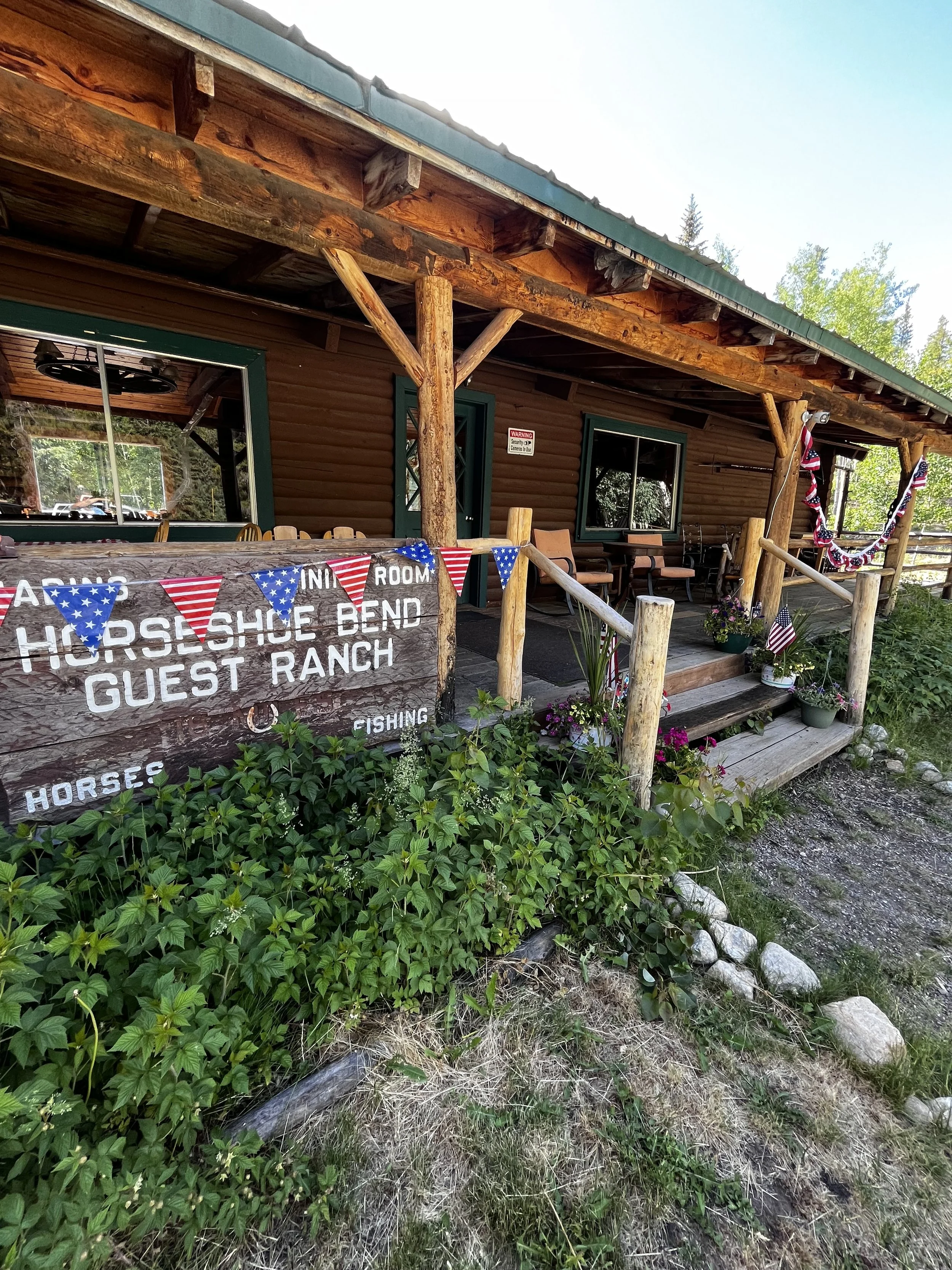 A rustic cabin with a porch decorated with American flags and bunting, surrounded by playfulness and flower pots, indicating a guest ranch called Horse Shoe Bend Guest Ranch.