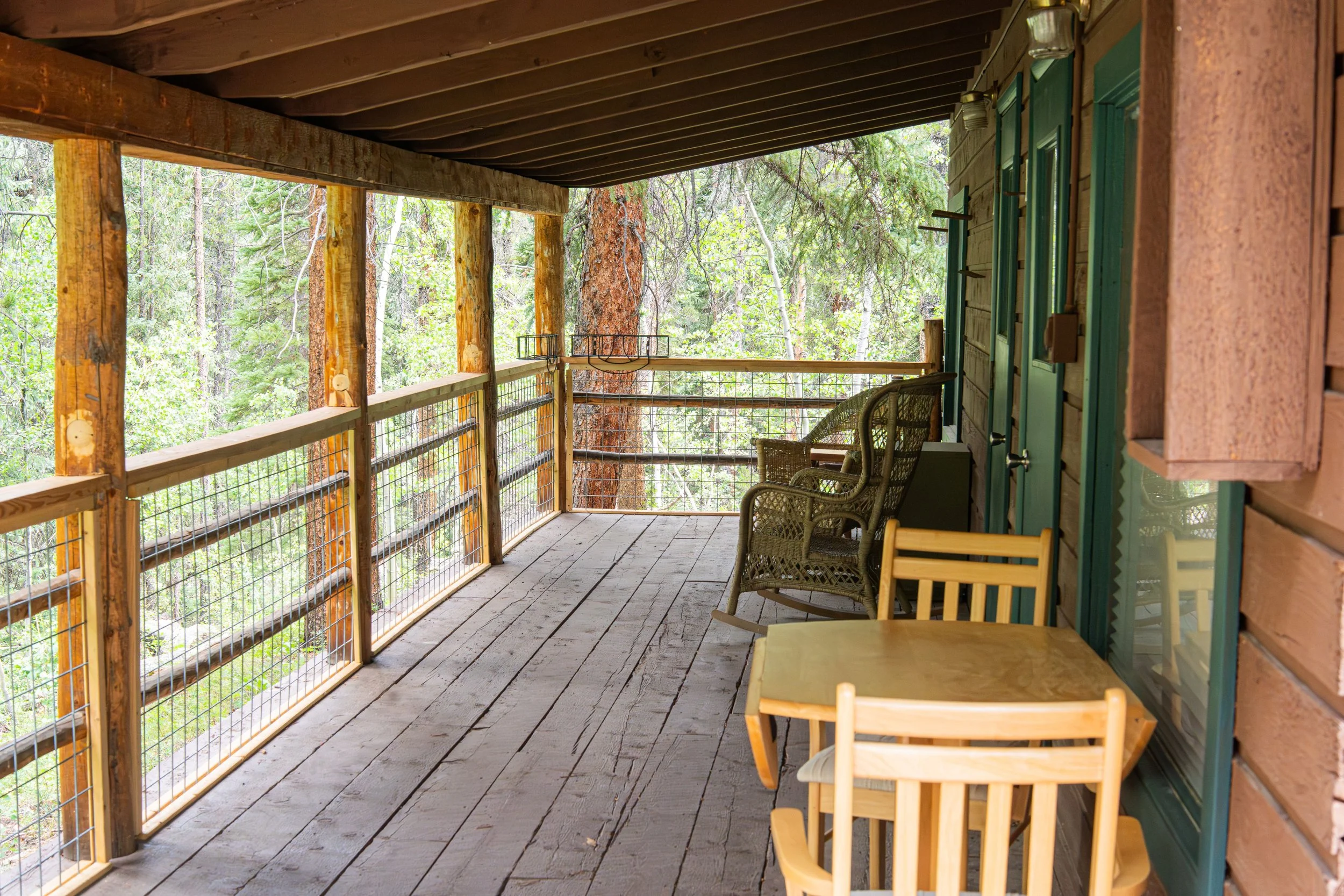 A rustic porch with wooden flooring and railing, surrounded by tall trees and greenery, featuring wicker and wooden chairs and a small wooden table.