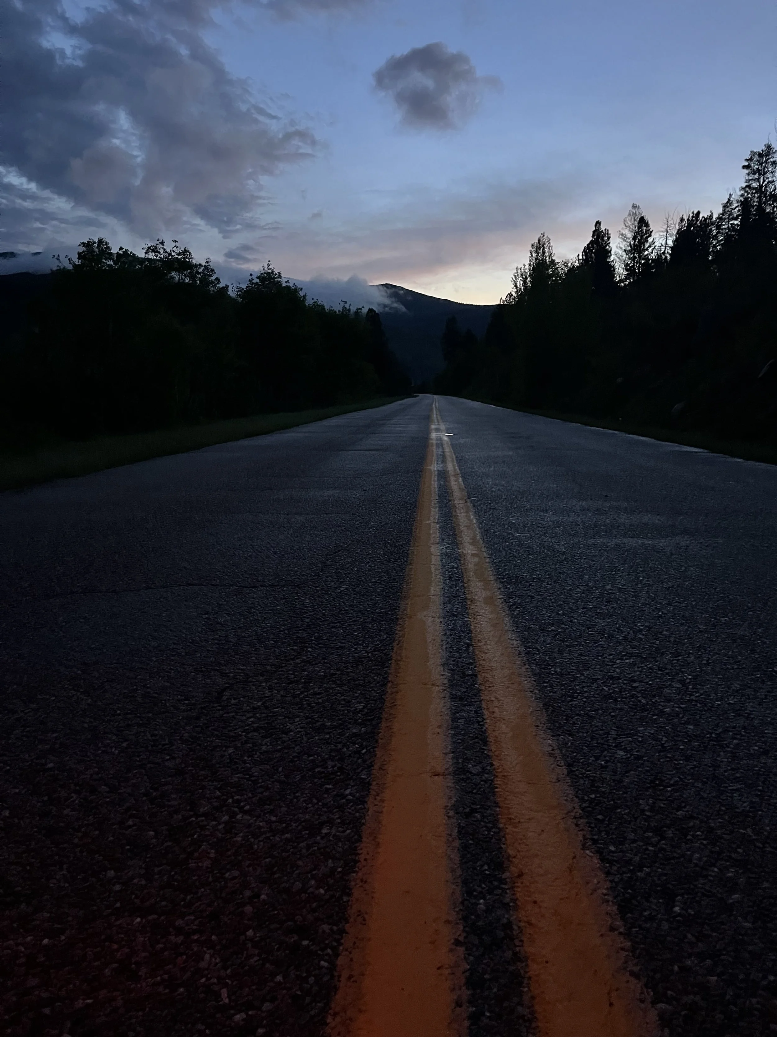 An empty two-lane road with yellow and black road markings, surrounded by trees and mountains, during dusk or dawn with a cloudy sky.