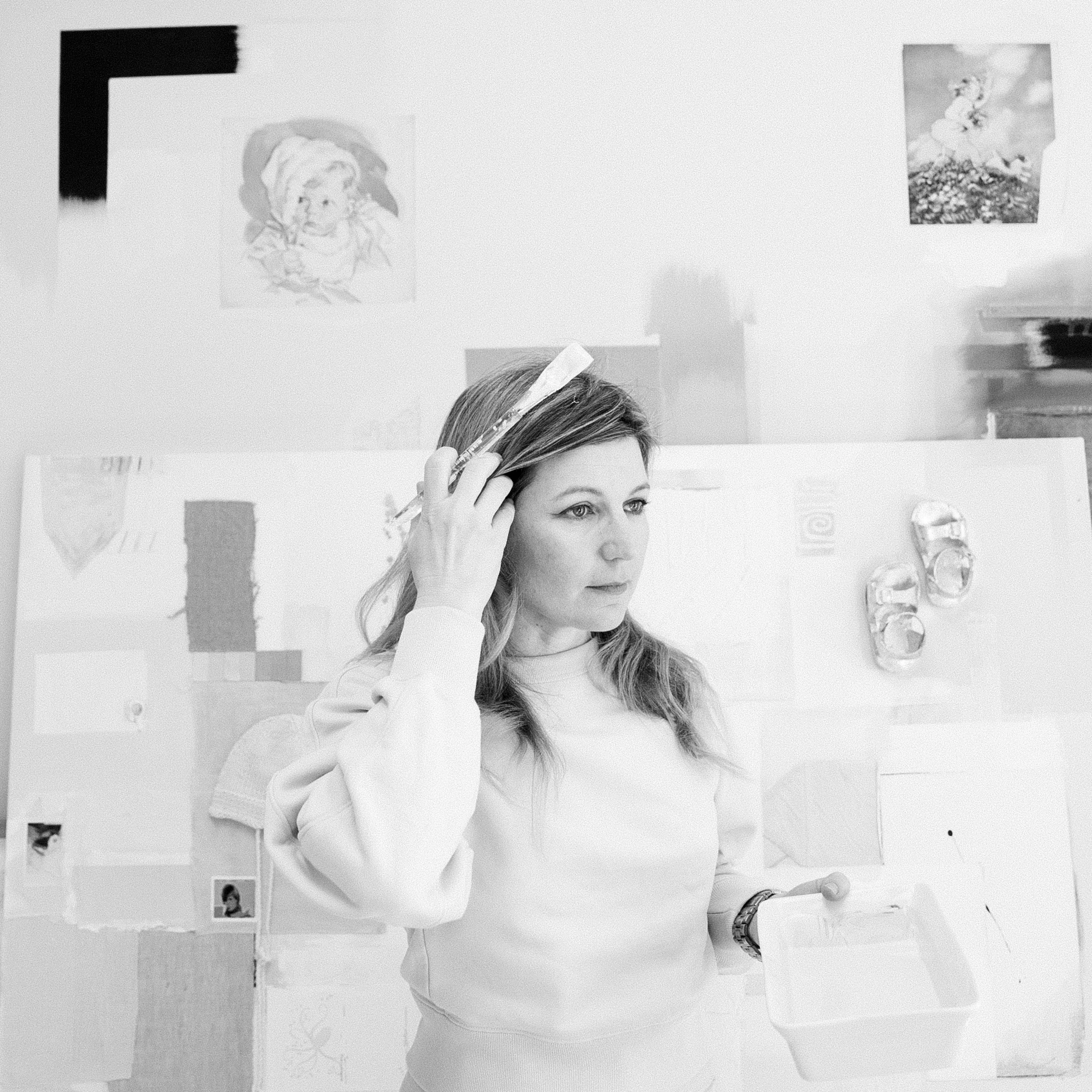 A woman with long hair, wearing a light-colored sweatshirt, stands in front of a wall decorated with various small pictures, including a child's portrait and an outdoor scene. She is holding a paintbrush to her forehead, looking thoughtful or focused.