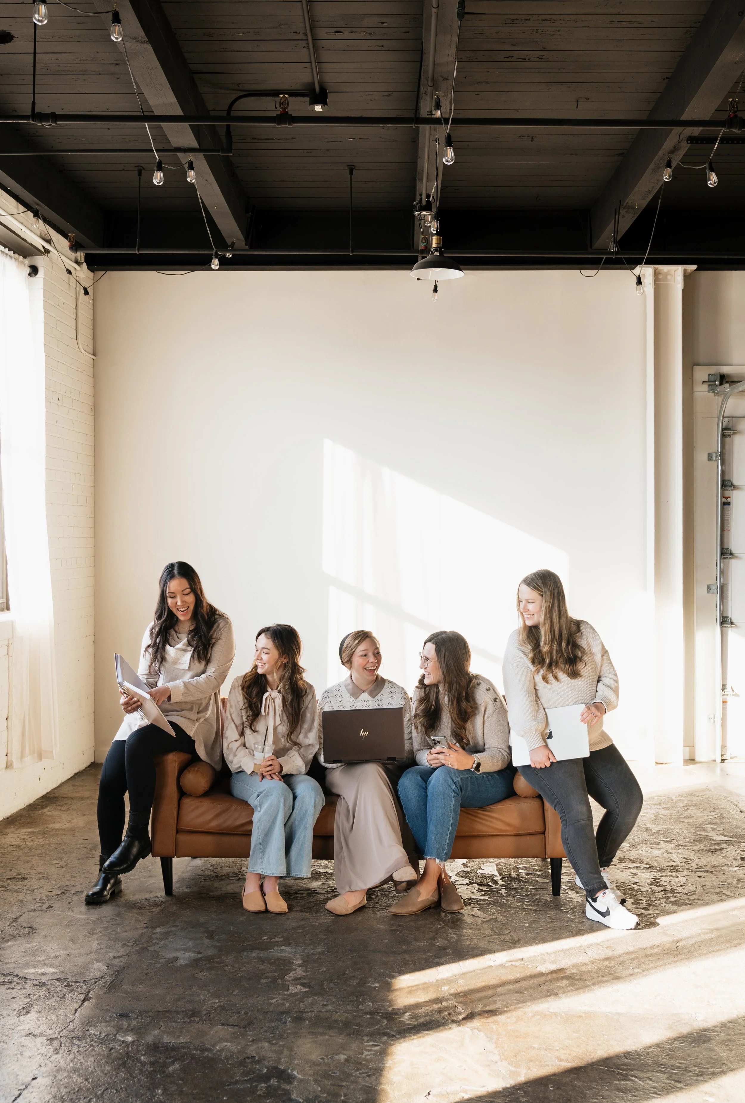 Five women sitting on a brown sofa in a bright room, engaged in conversation, with some holding papers and a laptop.